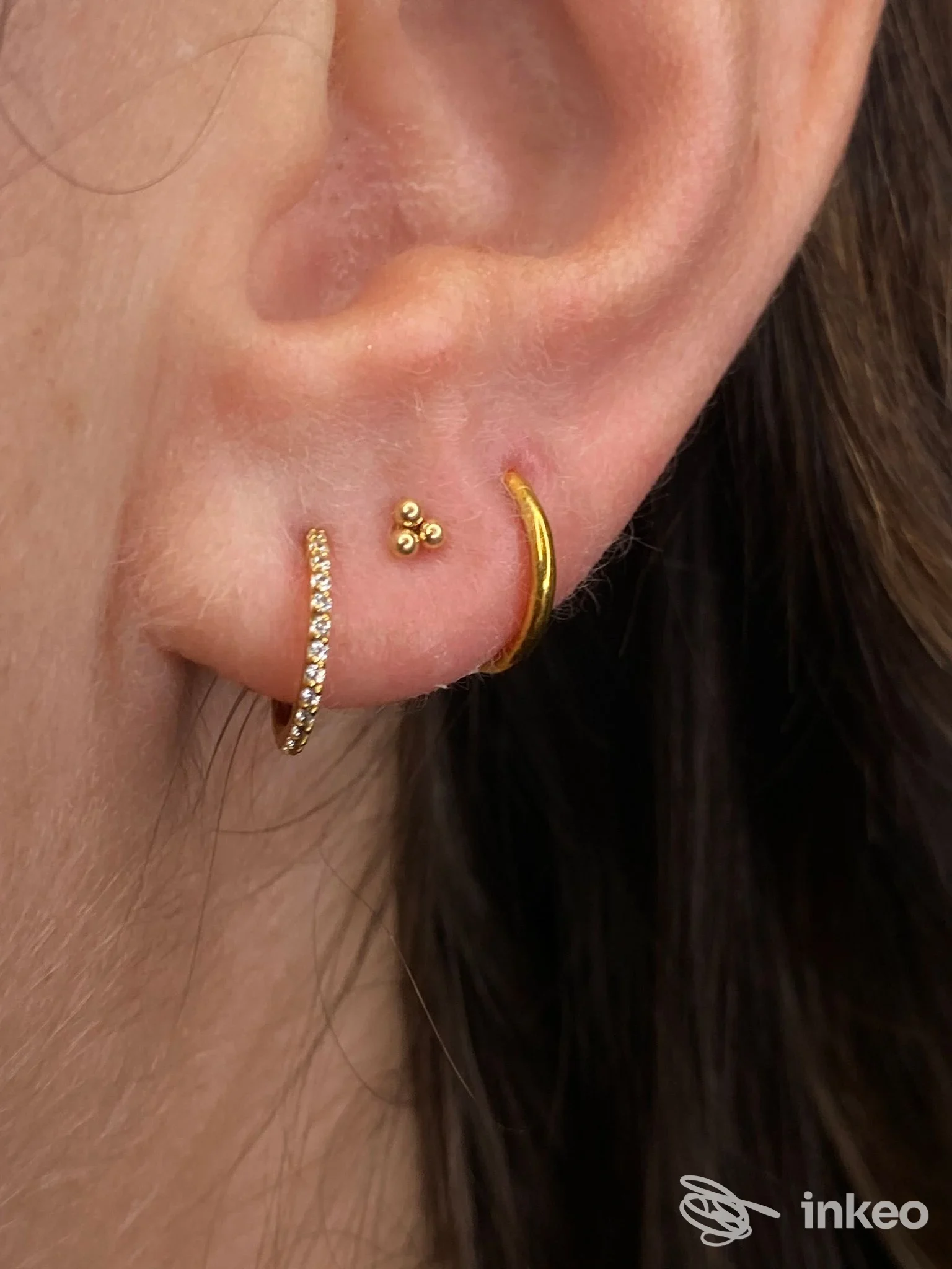 Orecchio sinistro con due piercing: un orecchino dorato a cerchio con micro-pietre e un piccolo perno, accompagnati da un terzo orecchino dorato sull’elica; nessun viso visibile.