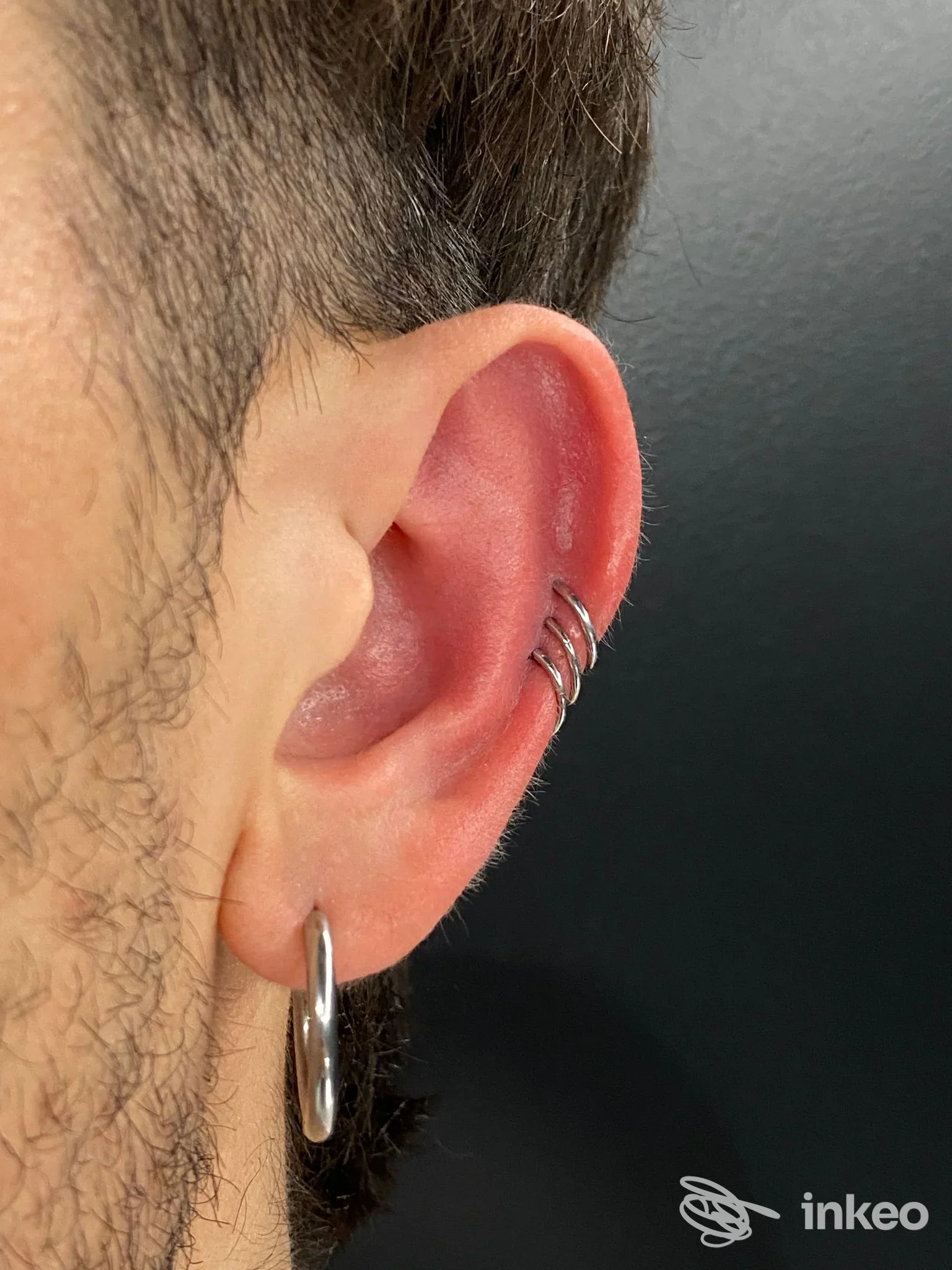 Orecchio sinistro con piercing multiplo sull’helix e orecchino al lobo, ritratto frontale parziale su sfondo scuro, senza testo visibile.