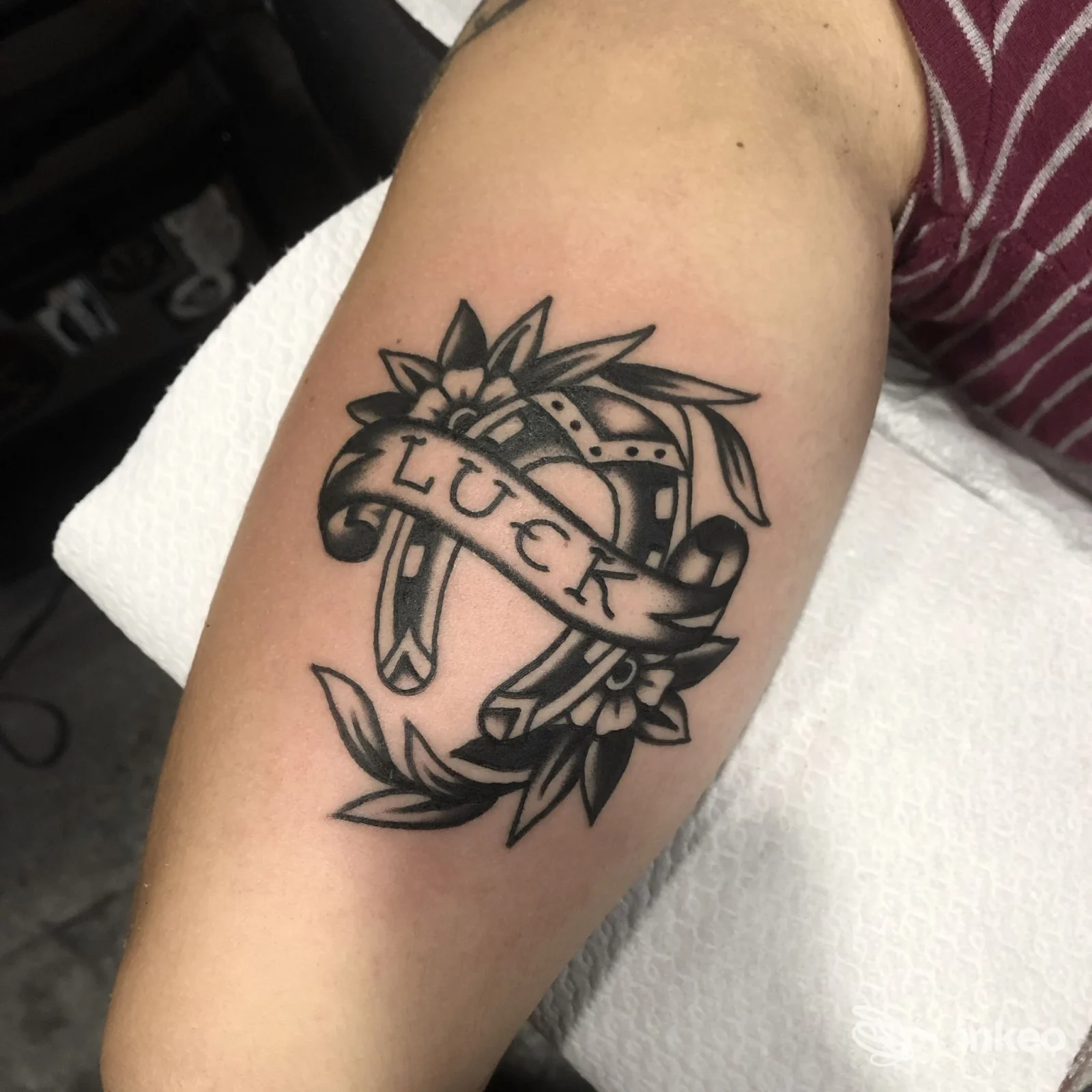 Tattoo sull'avambraccio: una cornice con fascia a bandiera contenente la scritta LUCK, decorazioni floreali che incorniciano il disegno, visibile una porzione di pelle.