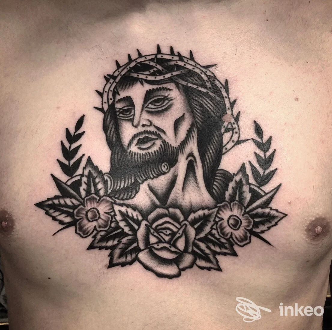 Tatuaggio sul torace raffigurante Gesù con corona di spine, barba e fiori ai lati, realizzato su pelle.