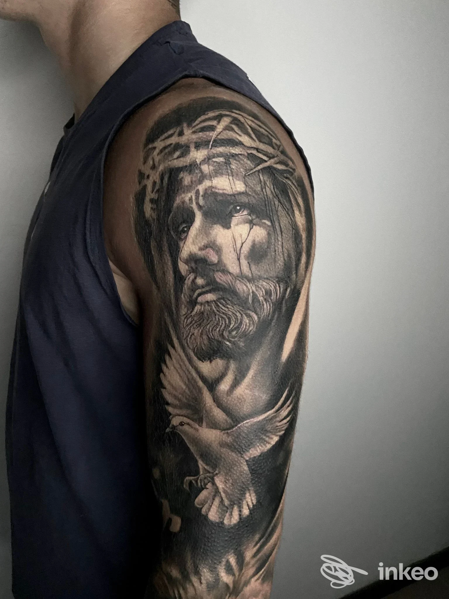 Tatuaggio realistico black & grey su spalla e bicipite con volto coronato di spine e colomba — Morrygan Tattoo Studio, Ferrara.