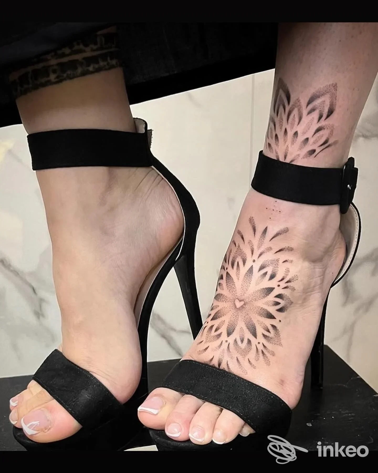 Tattoo floreale geometrico visibile su caviglia e polpaccio sinistri, realizzato sulla pelle con tonalità nere e grigi; sandali neri e sfondo neutro
