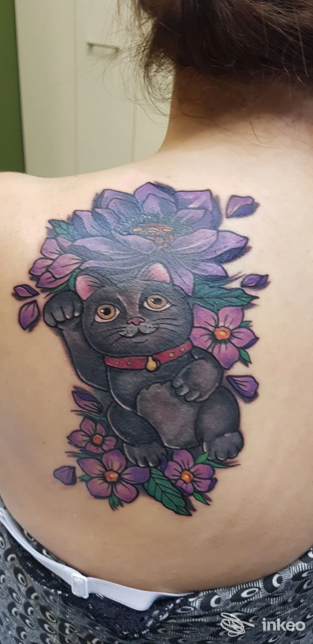 Tattoo sulla schiena raffigurante un gatto nero con collare rosso, circondato da grandi fiori viola e rosa, visibile nella parte superiore della schiena.