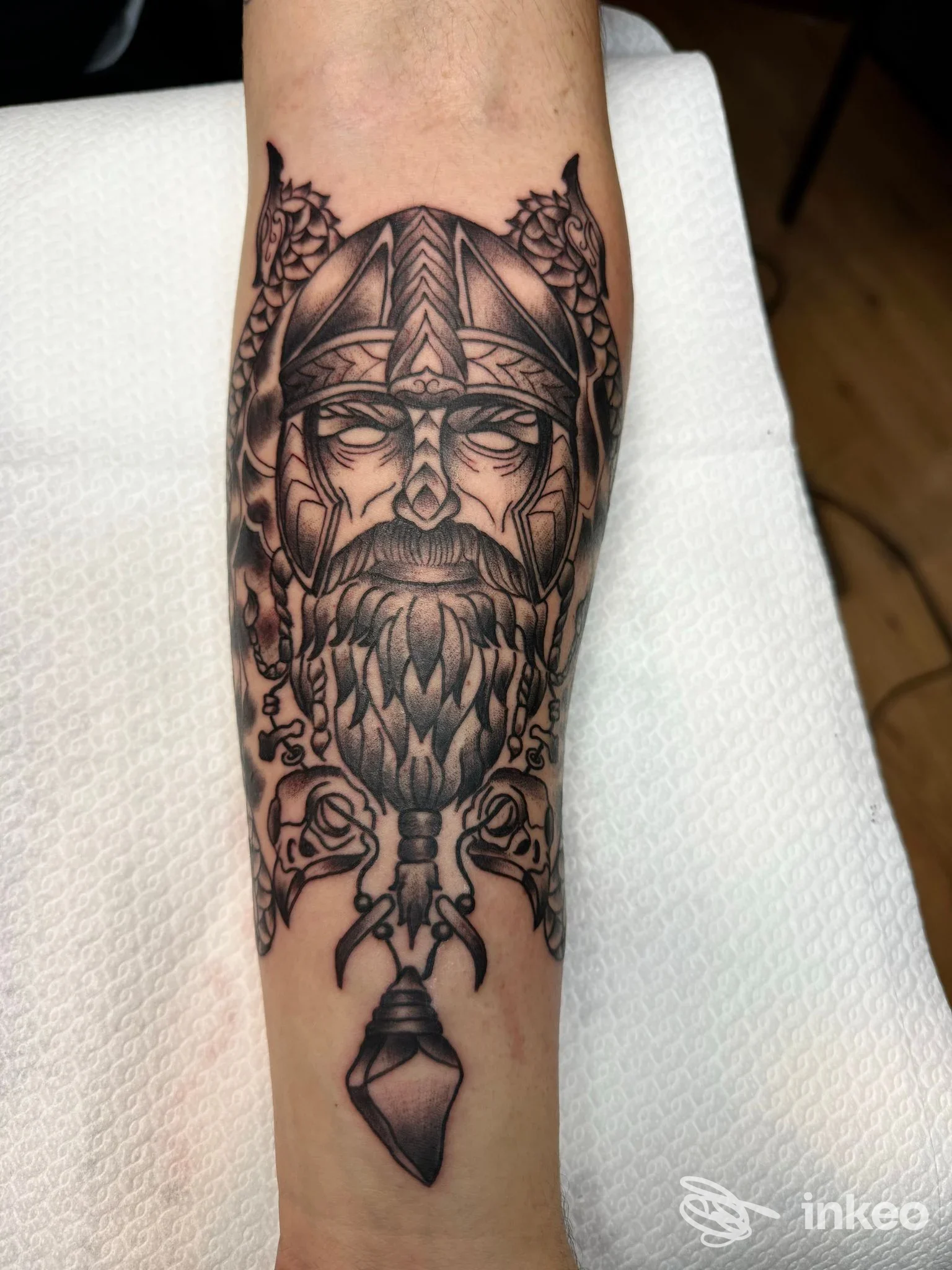 Tatuaggio in bianco e nero sull'avambraccio raffigurante un volto vichingo stilizzato con elmo, barba intrecciata e pendente, realizzato da Black Flag Tattoo a Riano.