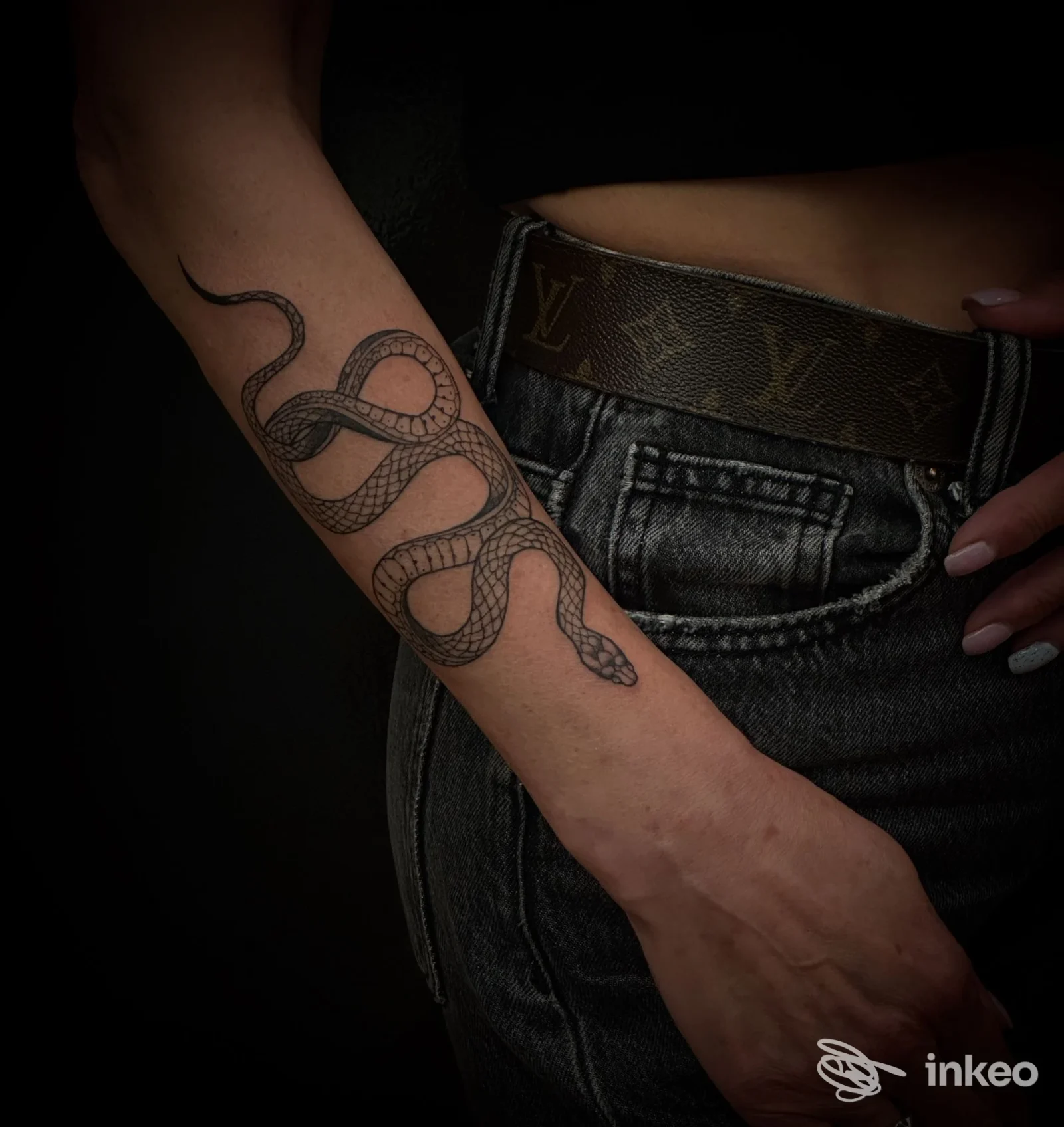 Tatuaggio raffigurante un serpente su avambraccio sinistro, realizzato sulla pelle, con sfondo scuro; indumenti denim e cintura visibili.