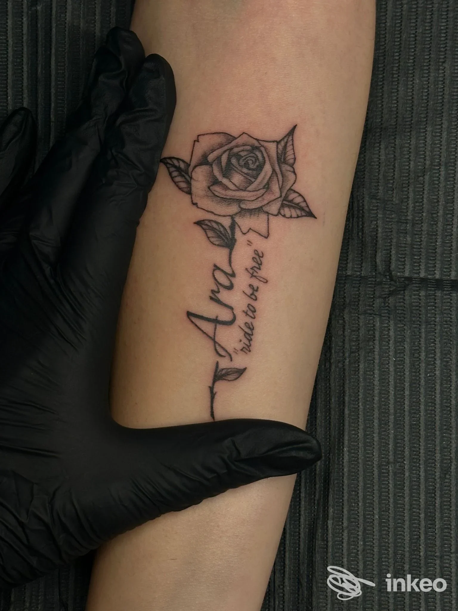 Tattoo realizzato su avambraccio sinistro: rosa stilizzata in alto e scritta corsiva lungo l’avambraccio, tenuto da una mano guantata nera.