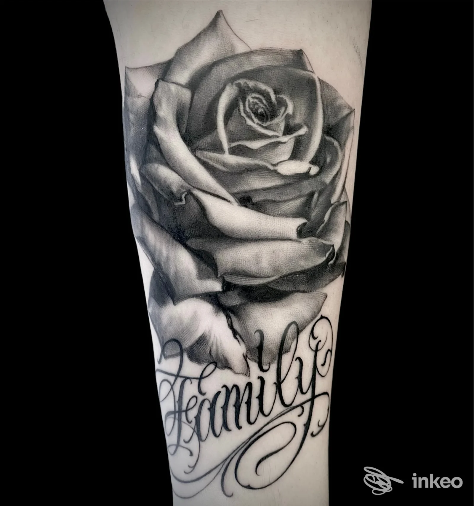 Tattoo realistico su avambraccio raffigurante una rosa dettagliata in bianco e nero con una scritta corsiva sottostante, stile sombre e sfumature complesse, eseguito su pelle umana.