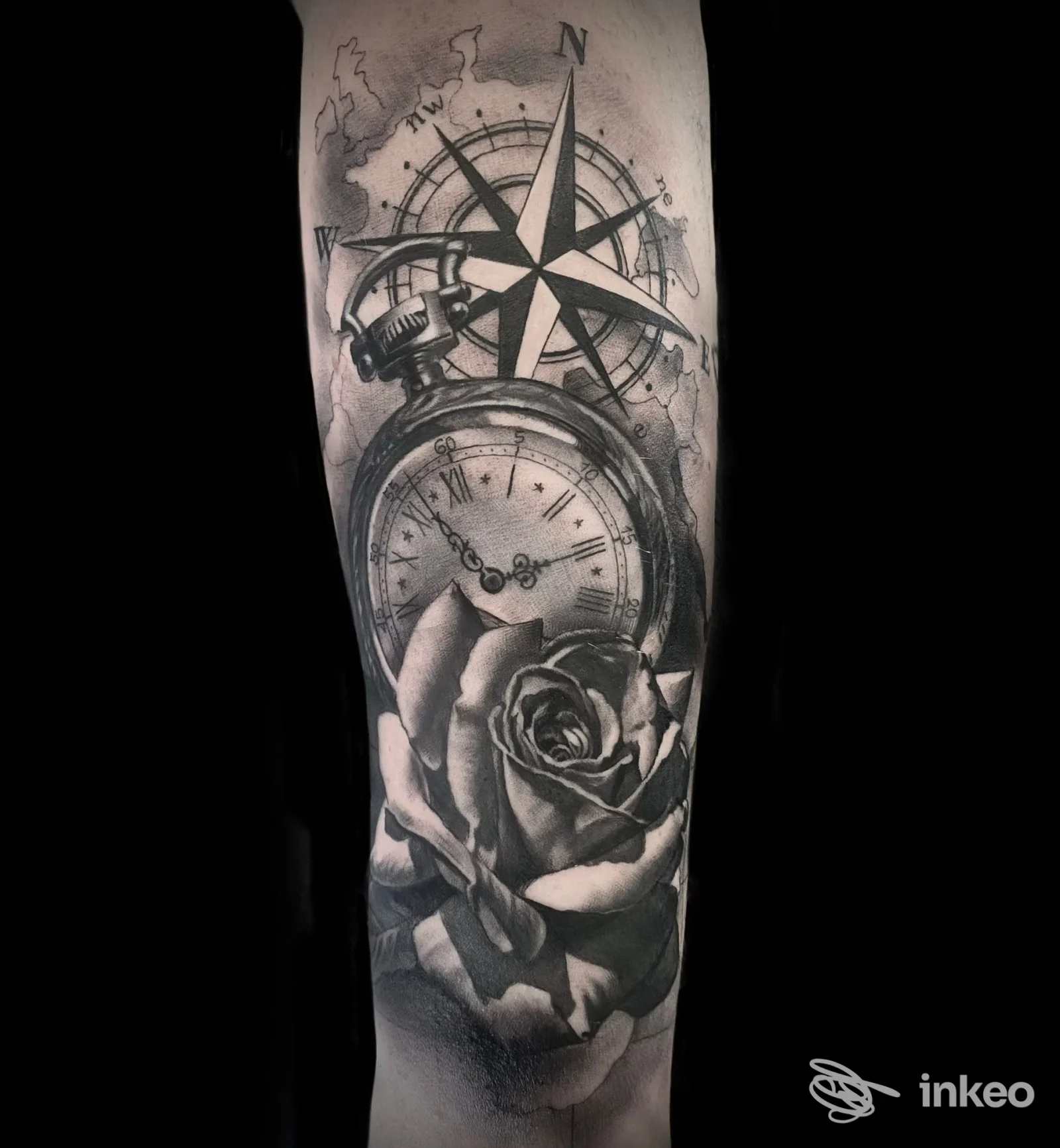 Tattoo sull'avambraccio raffigurante un orologio a tasca e una bussola con una rosa, in tonalità di grigio su sfondo scuro, realizzata sulla pelle.