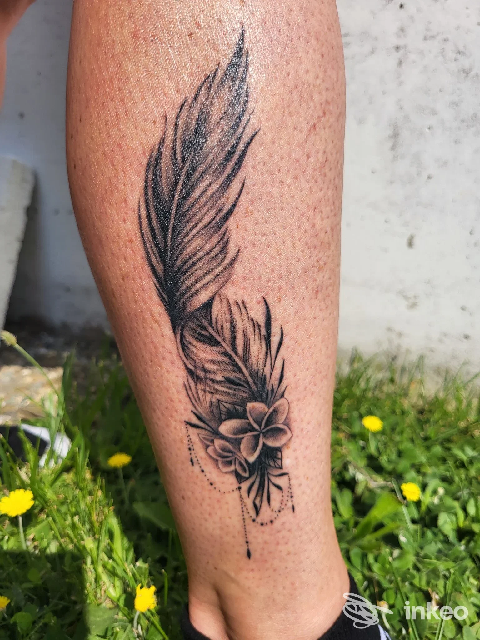 Tatuaggio a inchiostro nero e grigio a forma di piuma con fiori sul polpaccio, foto inviata da Inkwave Tattoo Lazise, Lazise.
