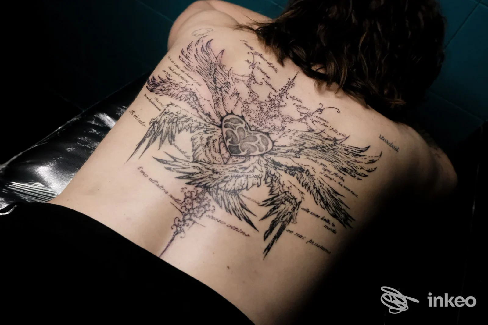 Tatuaggio grande sulla schiena raffigurante ali con cuore centrale, disegno stilizzato con linee decorative, visibile su pelle reale durante una sessione in studio.