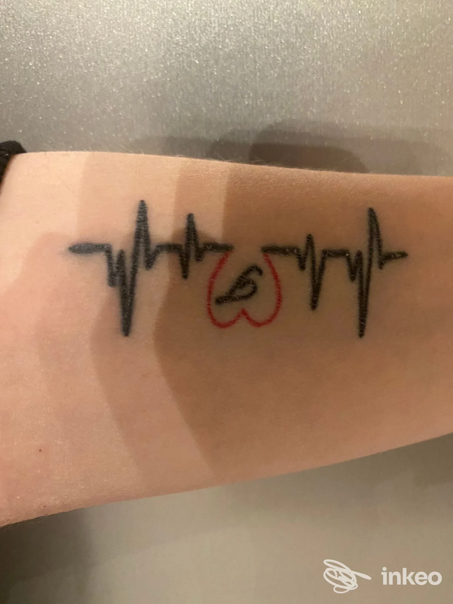 Tatuaggio sull'avambraccio: linea ECG nera con cuore rosso al centro, senza testo o elementi visivi esterni visibili.