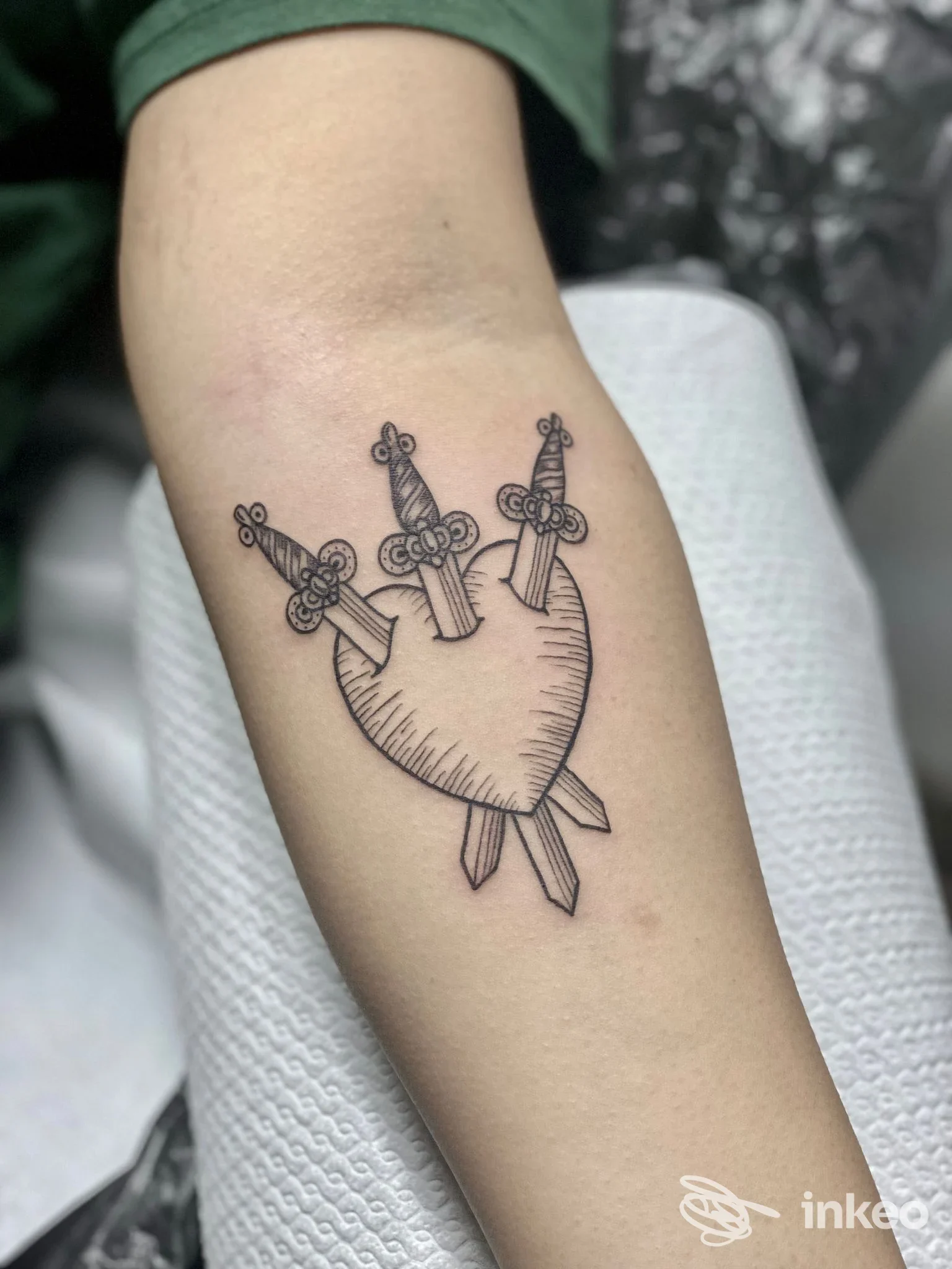 Tatuaggio linework nero di un cuore trafitto da tre pugnali sull'avambraccio, fotografato presso Downtown Ink Verona.