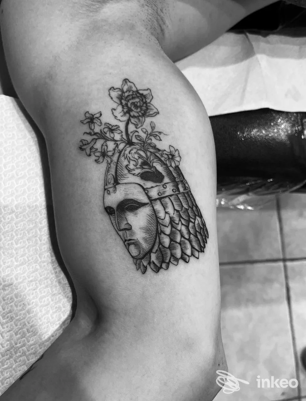 Tatuaggio a inchiostro nero su interno braccio raffigurante una maschera con elmo e fiori, Tattoon Studio, Bitonto.