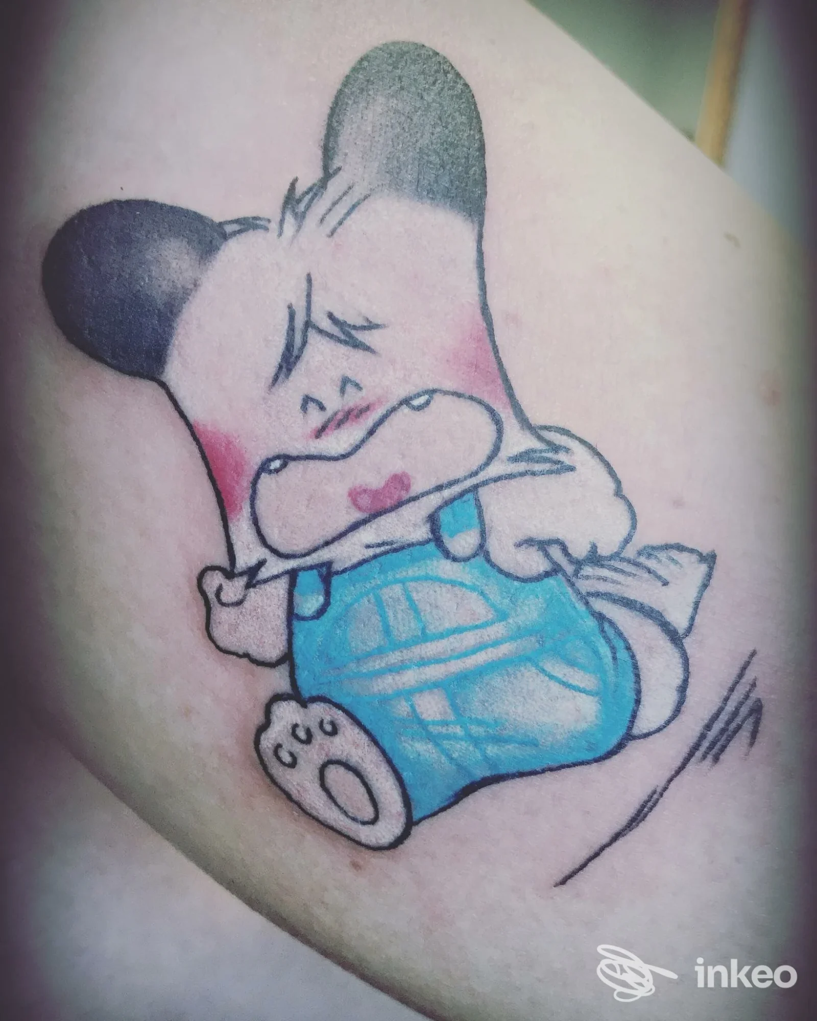 Tatuaggio cartoon su avambraccio: personaggio con salopette azzurra, guance rosa e dettagli neri — Fata Morgana Studio Tattoo, Agliana.