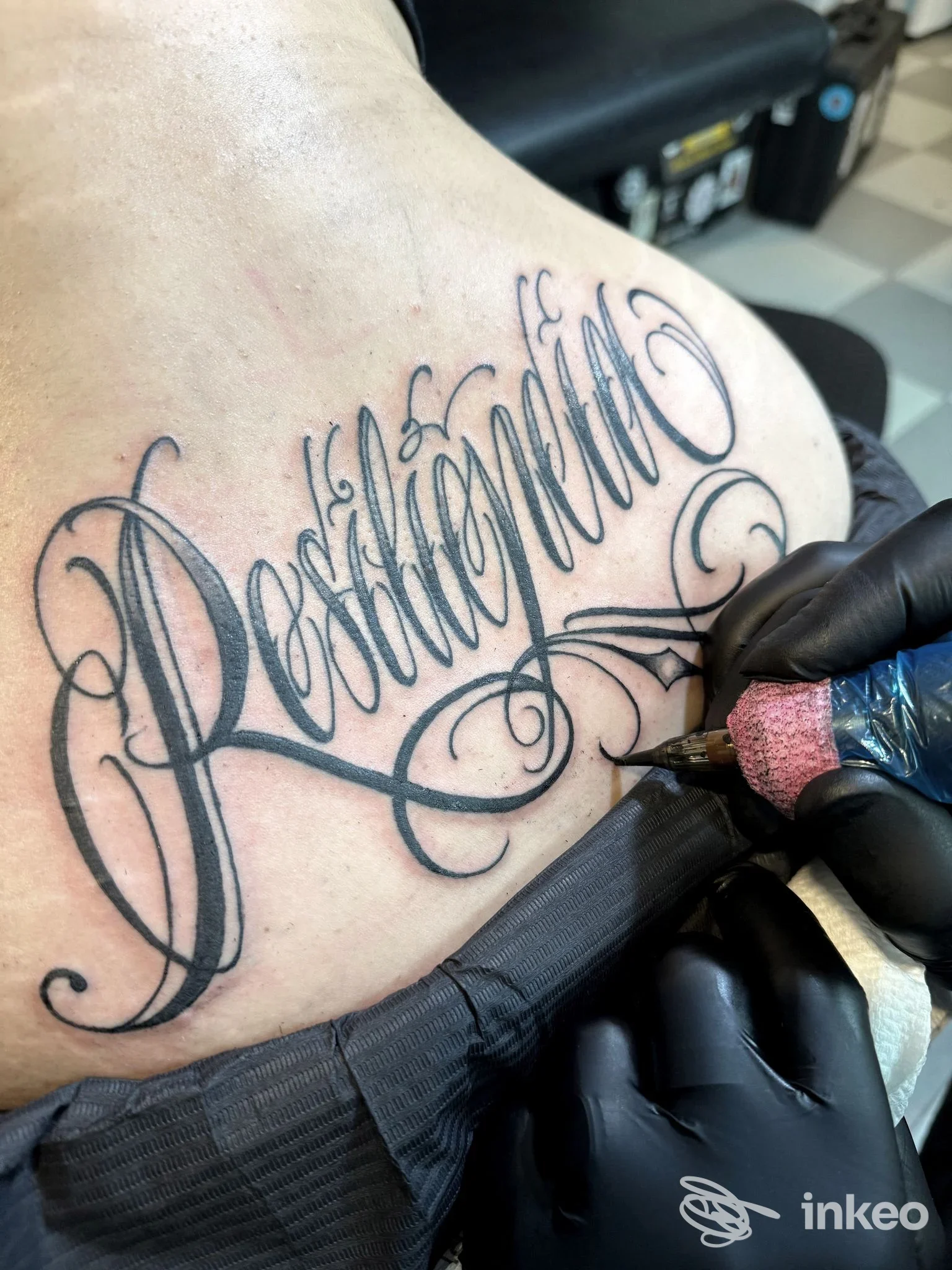 Tatuaggio lettering nero in corso sulla spalla, fotografato nello studio Pagan Tattoo Art Studio a Latina.