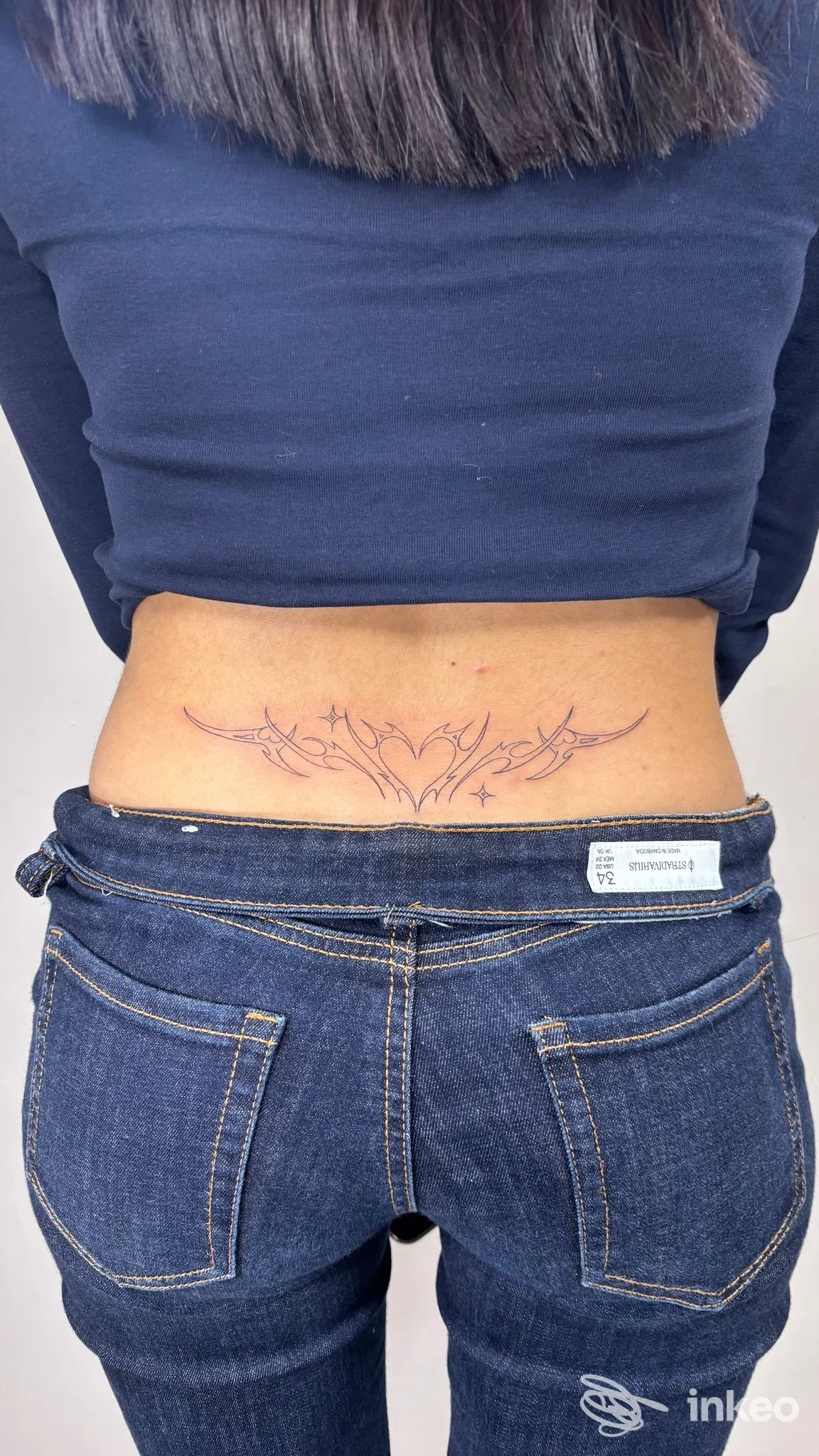 Tatuaggio ornamentale lineare sul busto inferiore della schiena di una persona vista di schiena, senza volto visibile; maglia blu sollevata e jeans.