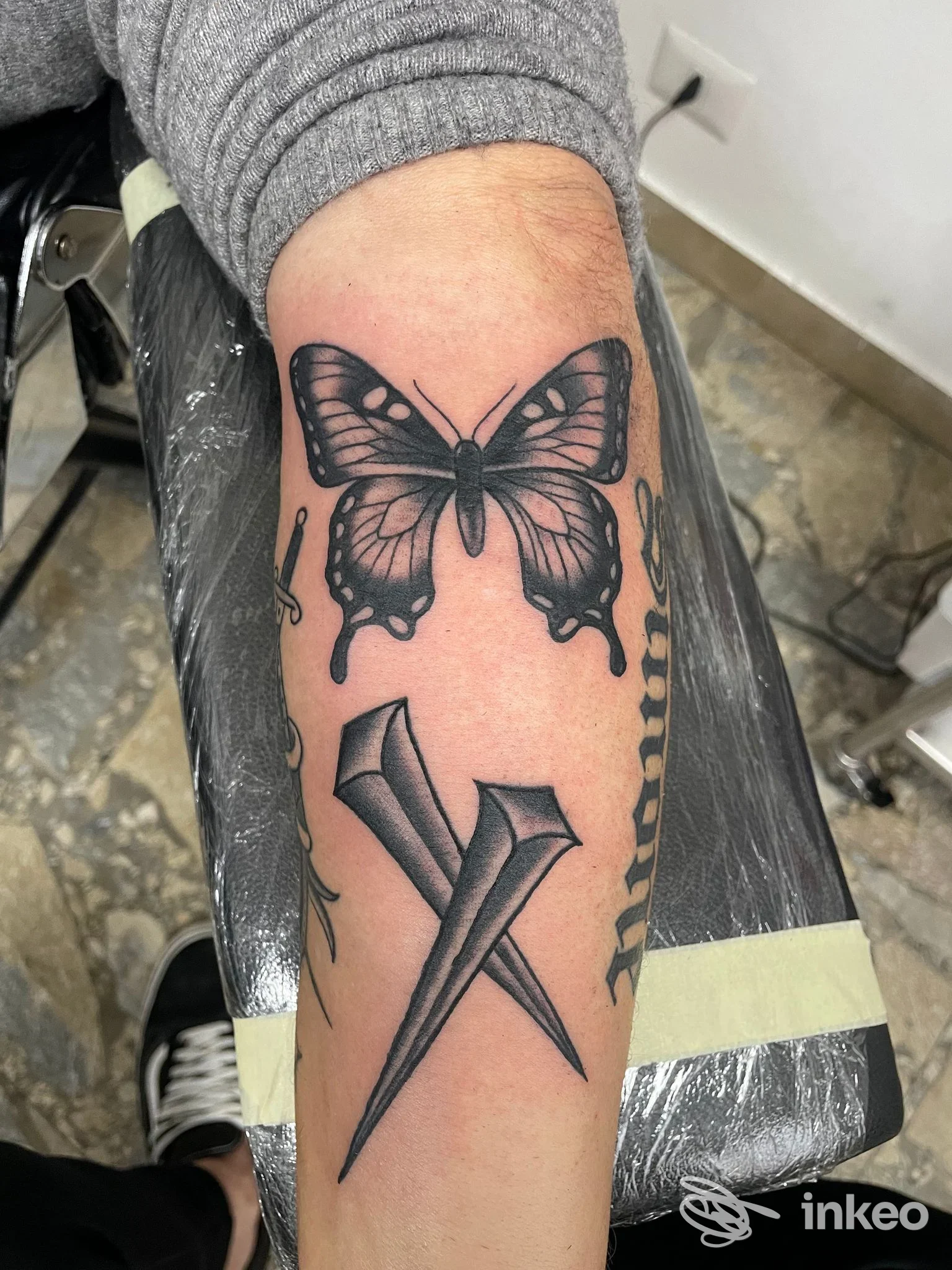 Tatuaggio farfalla e chiodi incrociati in bianco e nero sull'avambraccio, fotografia dallo studio Inked Up Tattoo Shop, Sabaudia.