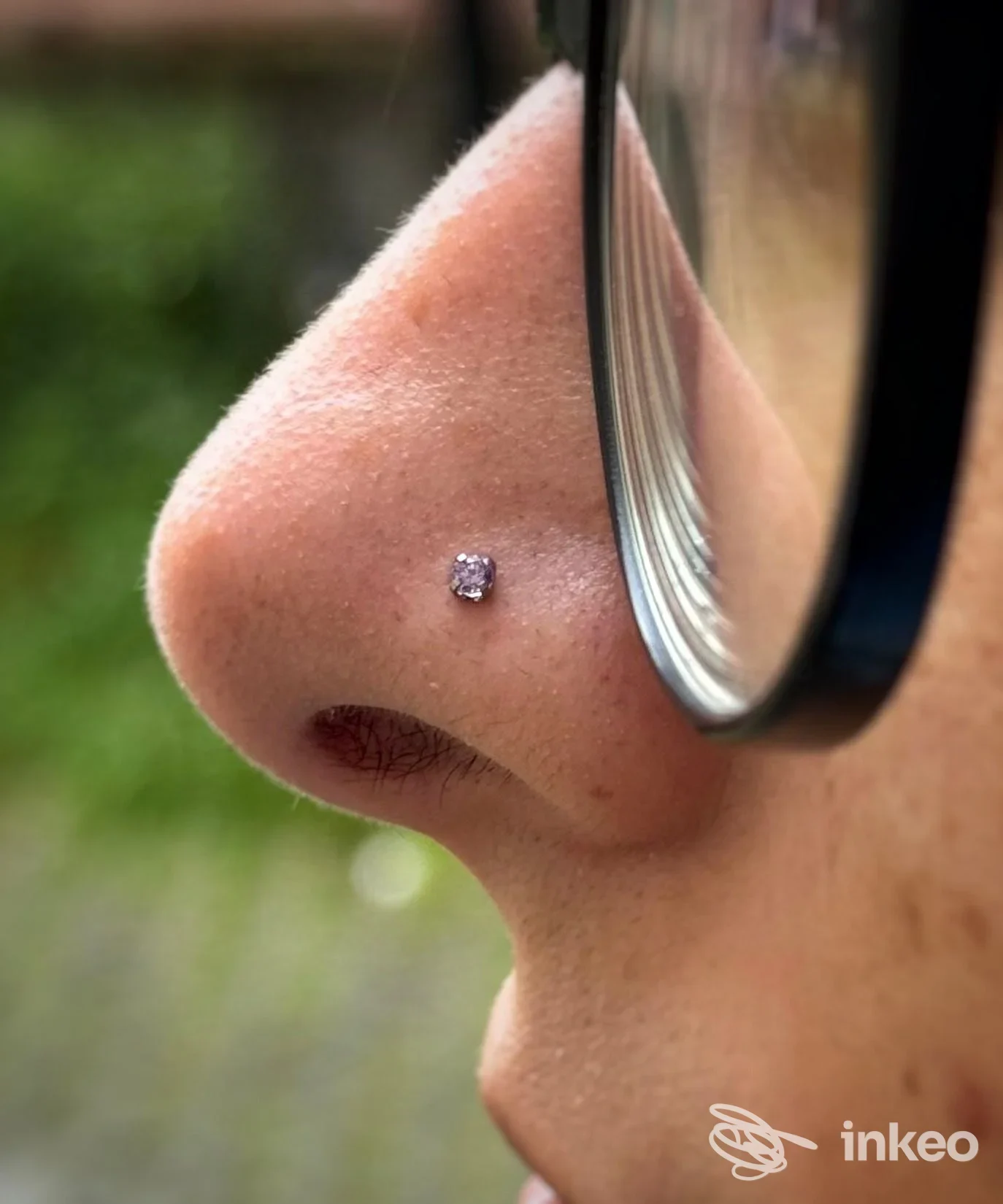 Primo piano di una persona con un piercing al naso molto piccolo e lucente, visibile accanto agli occhiali; realizzato direttamente sulla pelle, sotto un viso parziale.