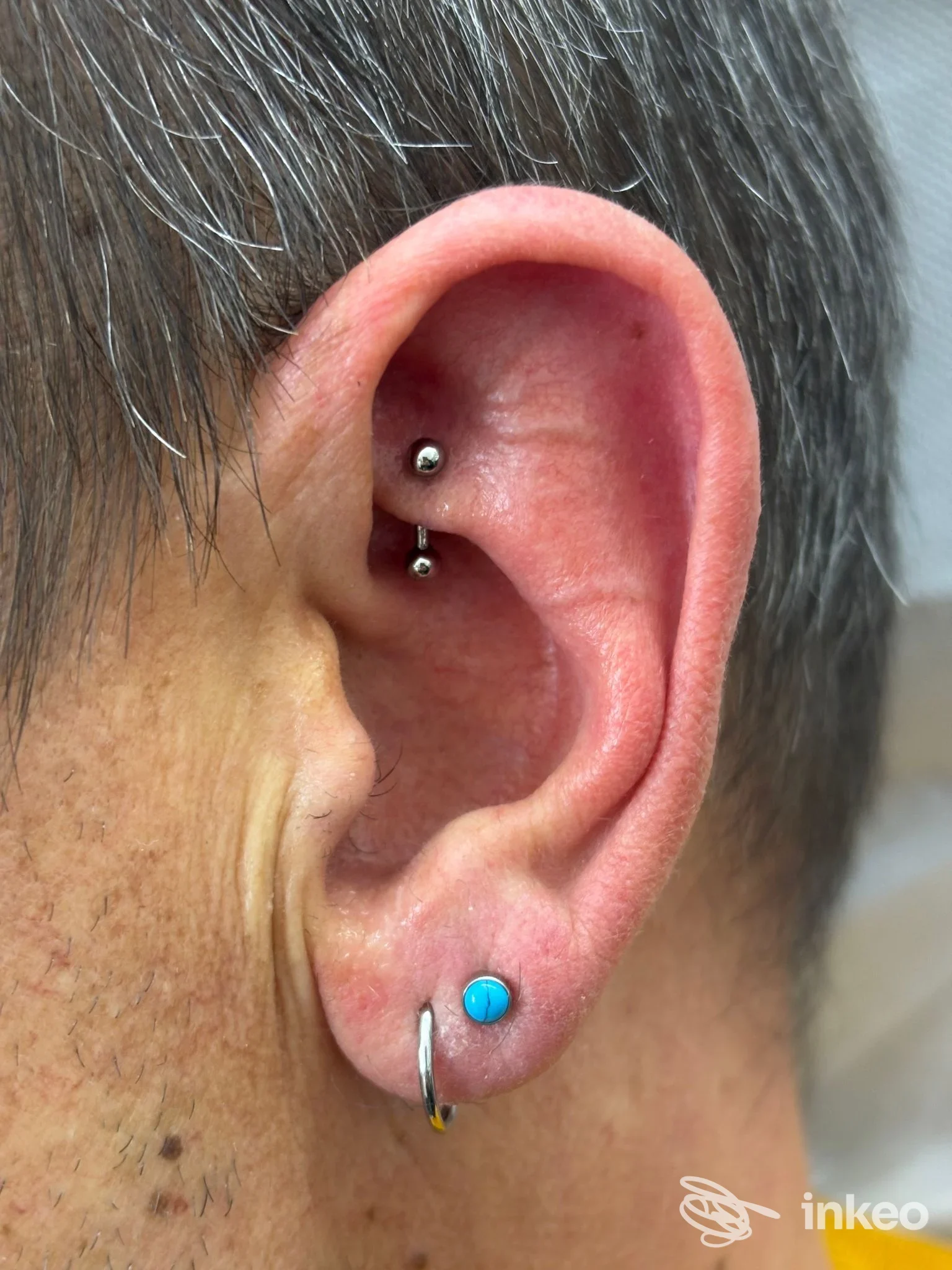 Piercing multipli all'orecchio visibili: due piccoli piercings sull'helix e un cerchio al lobo, con pallino azzurro, sul lato sinistro dell'orecchio. Studio Bp Barbara Bordin.
