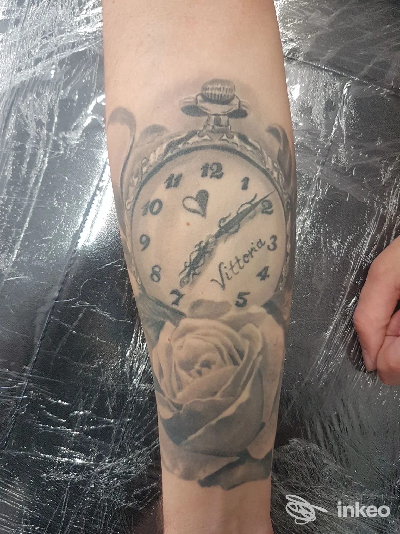 Avambraccio con tatuaggio realistico di orologio da tasca e rosa, elegante ornamento in tonalità nere e grigie, nessun volto visibile, sfondo lucido.