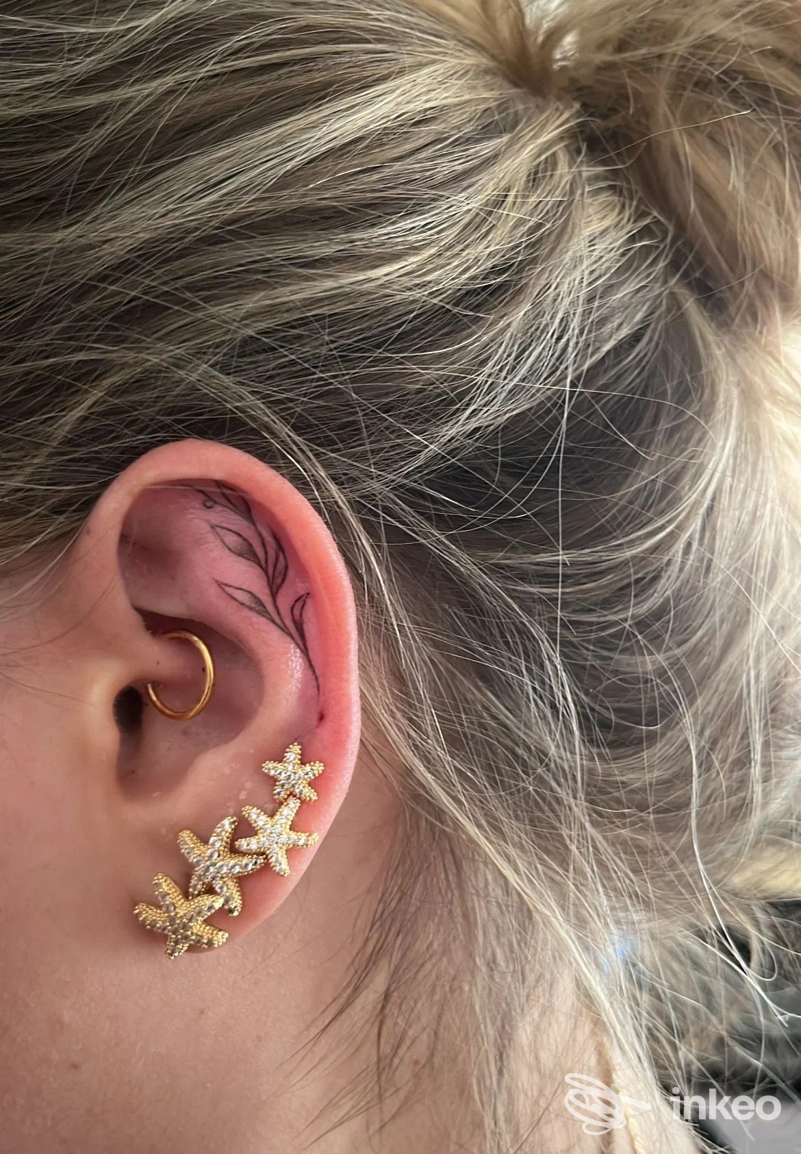 Dettaglio di orecchio con tatuaggio a rametto sulla cartilagine e orecchini dorati, foto fornita da WhyNot_Ink a Castel d'Azzano.
