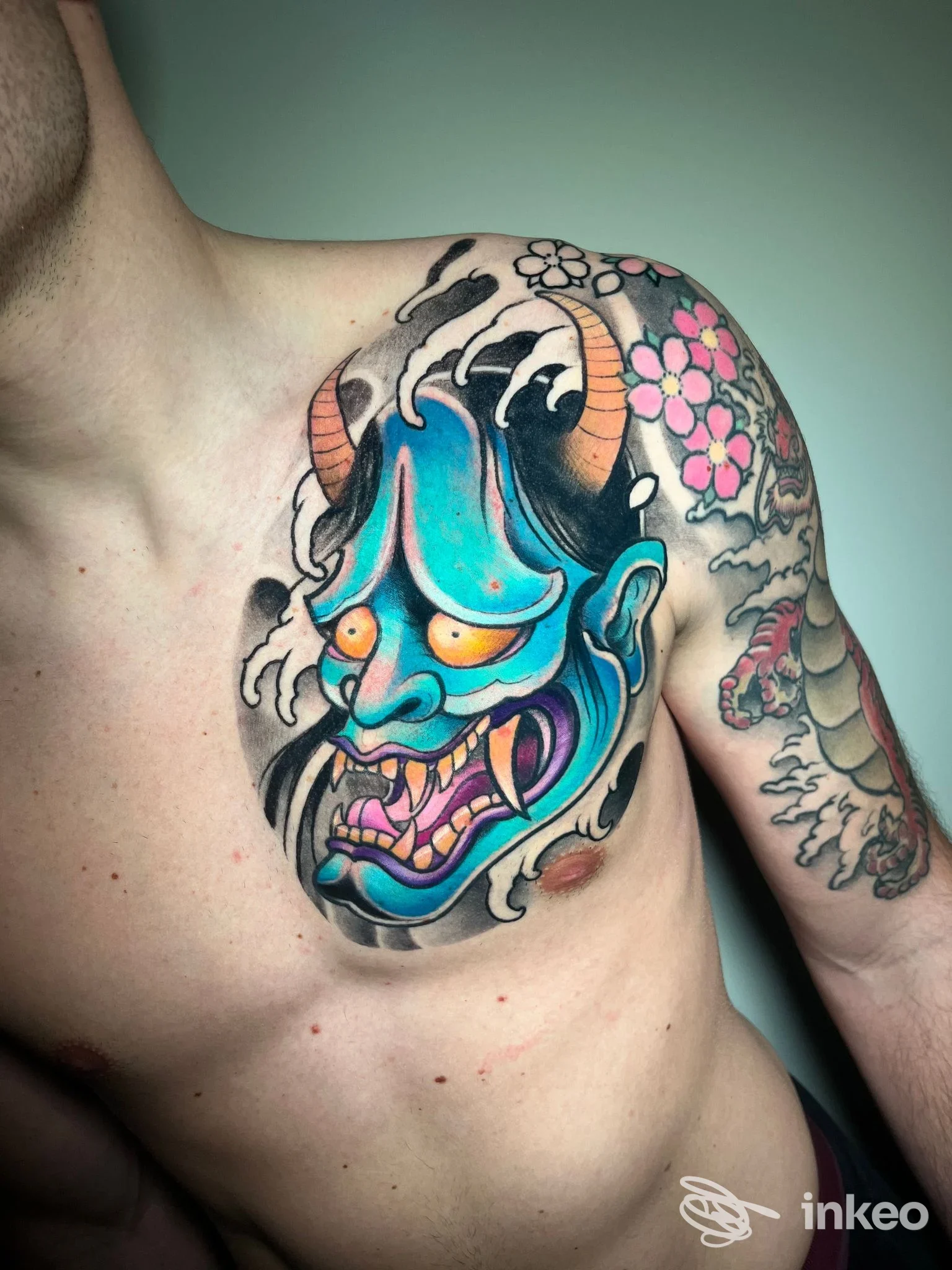 Tattoo a colori su spalla e petto raffigurante una maschera demoniaca blu con corna, accompagnata da fiori rosa sul braccio; dettaglio nitido e ben definito sulla pelle.