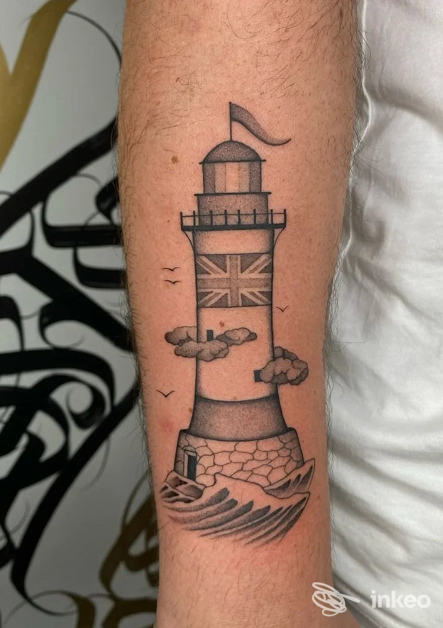Tattoo su avambraccio raffigurante un faro con bandiera britannica sul fusto, nuvole e onde ai piedi, eseguito in stile linework nero.