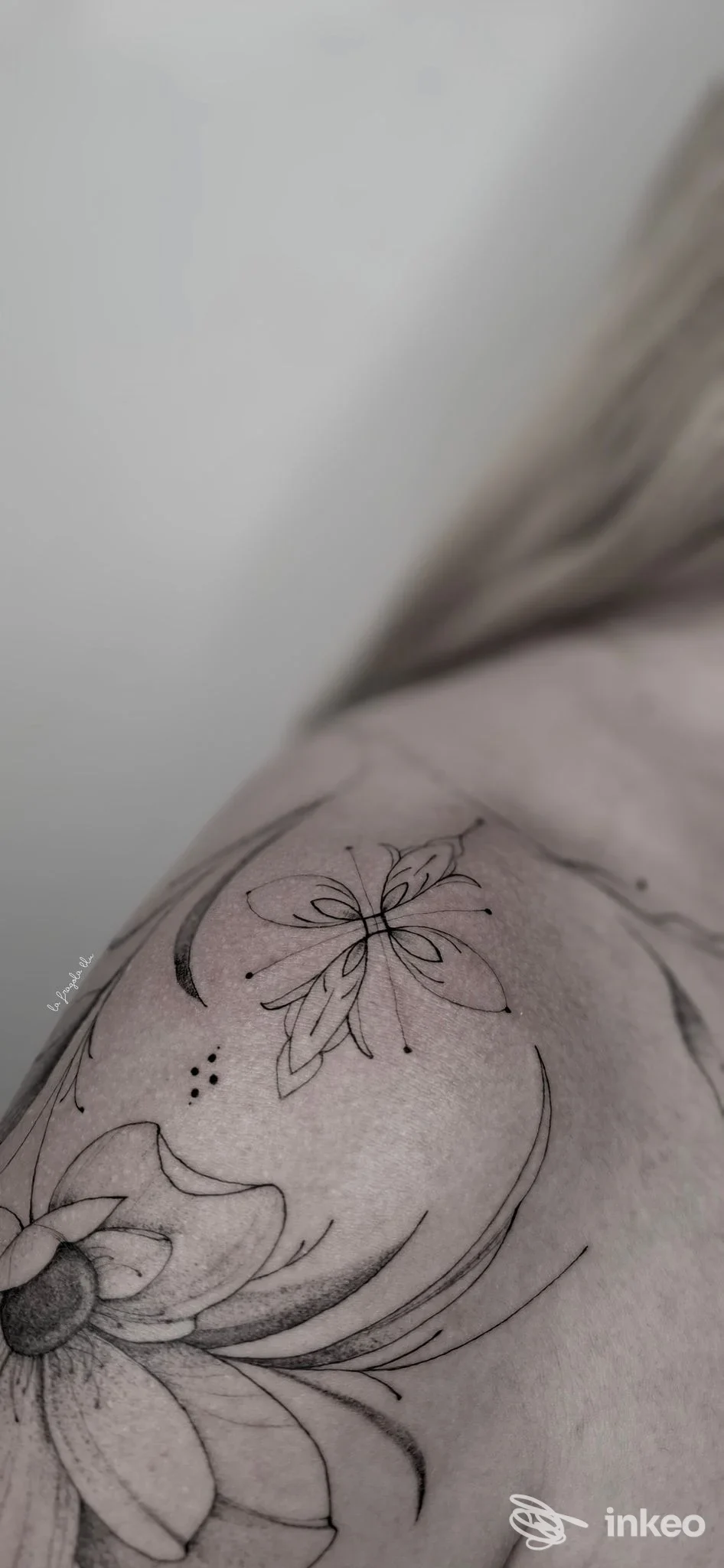 Tattoo floreale su pelle visibile su spalla, linee nere sottili e dettagli delicati. Nessun volto visibile.