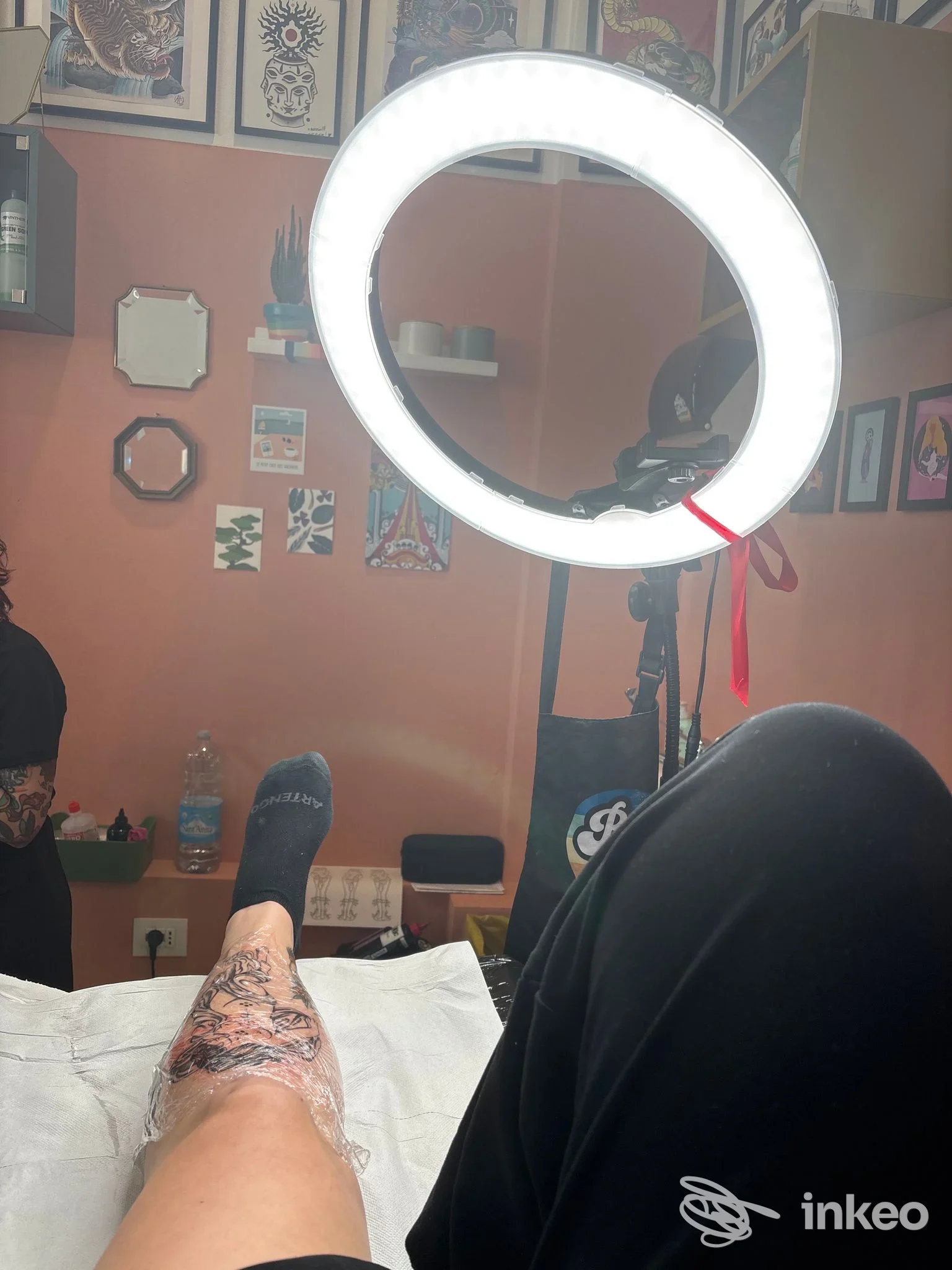 Foto in studio di Belly Button Tattoo Studio, Calenzano: tatuaggio appena fatto sul polpaccio di una persona, area coperta da plastica protettiva, lampada ad anello visibile.