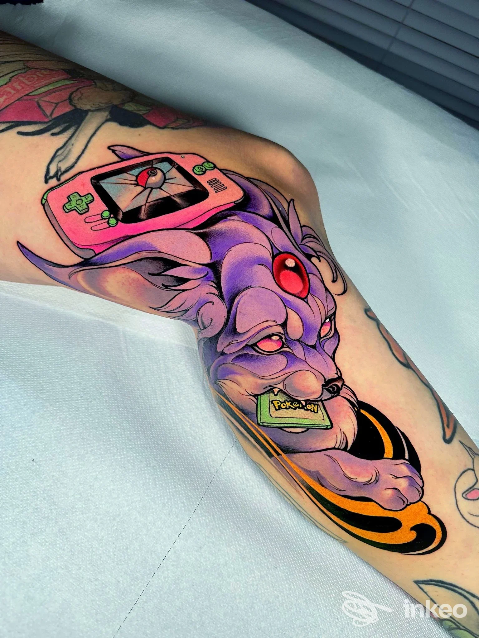 Tatuaggio colorato su gamba: creatura viola con console rosa, dettagli gialli e rossi, eseguito sulla gamba inferiore, sfondo di telo azzurro in studio.