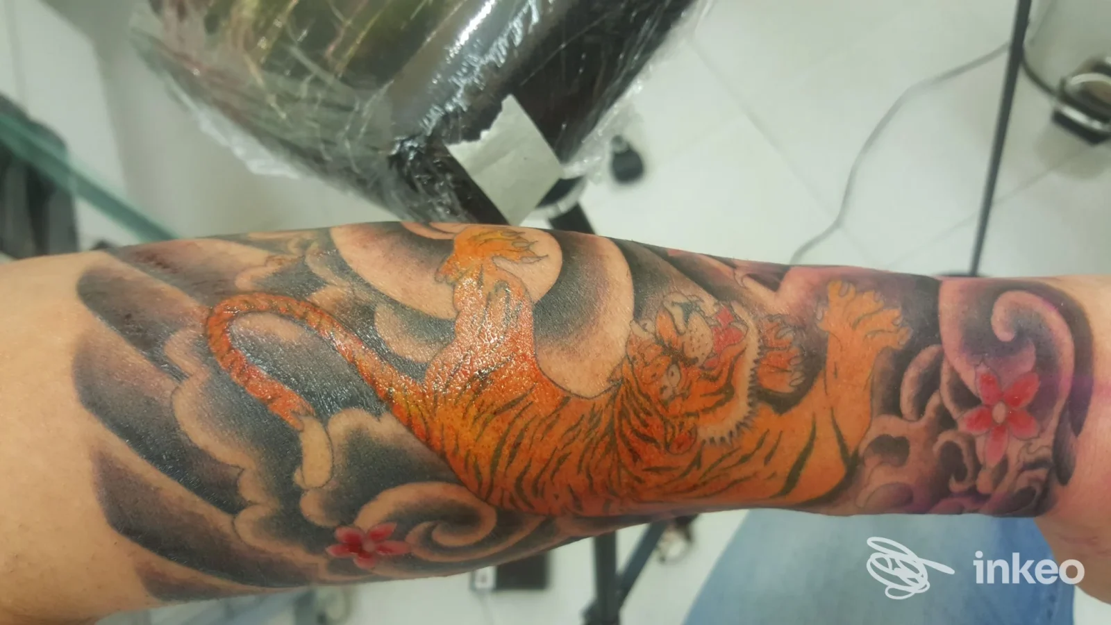 Tatuaggio realistico di tigre arancione sul braccio, sfumato con nuvole e fiorellini rosa, visibile lungo l'avambraccio.