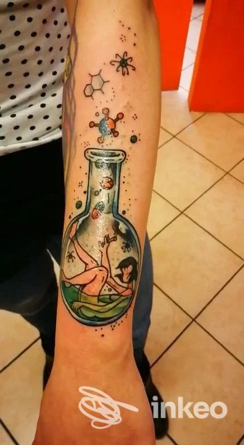 Tatuaggio colorato sull'avambraccio raffigurante una donna in una boccetta con bolle e molecole, realizzato da Sons Of Ink a Oria.