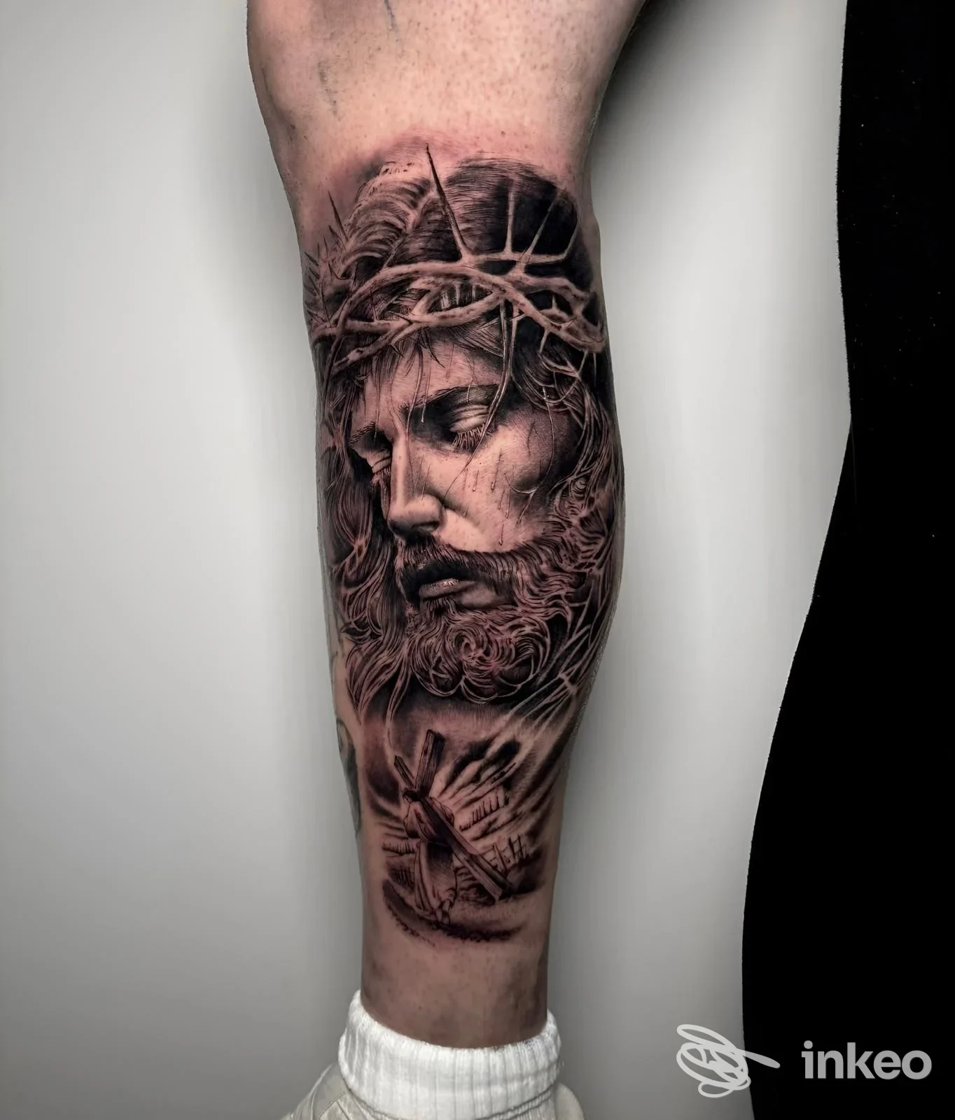 Tatuaggio realistico sul polpaccio raffigurante il volto di Cristo con corona di spine e mani giunte, foto fornita da Indelebile Studio Tattoo - Tatuaggi Sava, Sava.