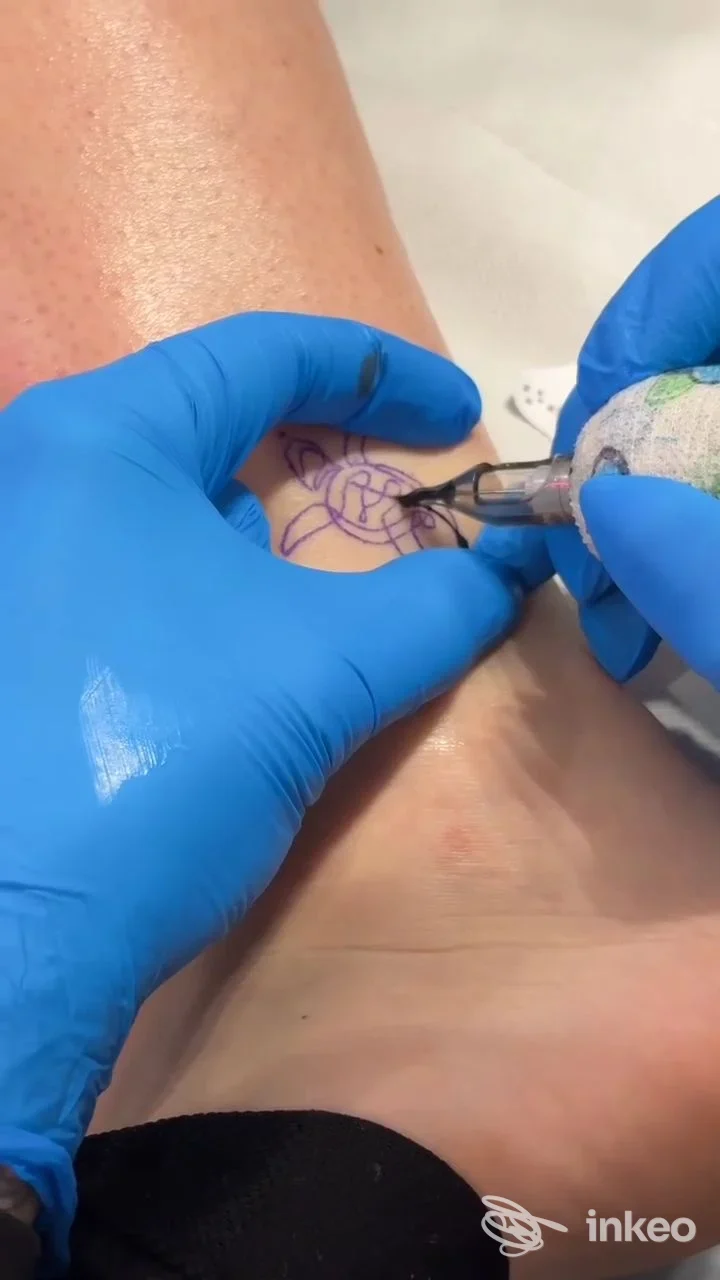 Procedura di tatuaggio sul collo del piede eseguita da SaraMau a Montemurlo, mani con guanti e macchinetta in azione sullo stencil.