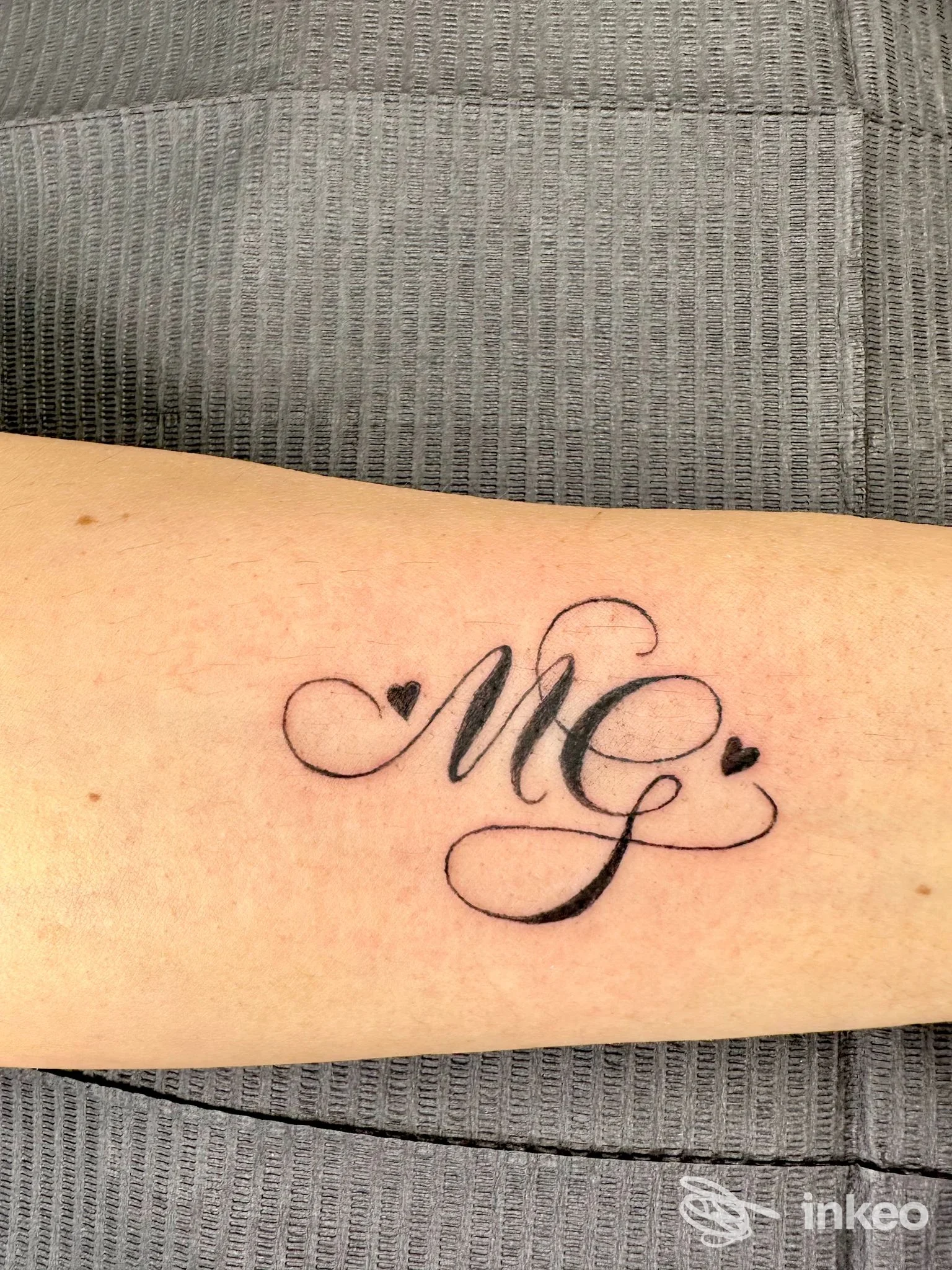 Tatuaggio con iniziali corsive 'MG' e due cuoricini sull'avambraccio, lavoro di Lucilla Panerai a Calenzano.