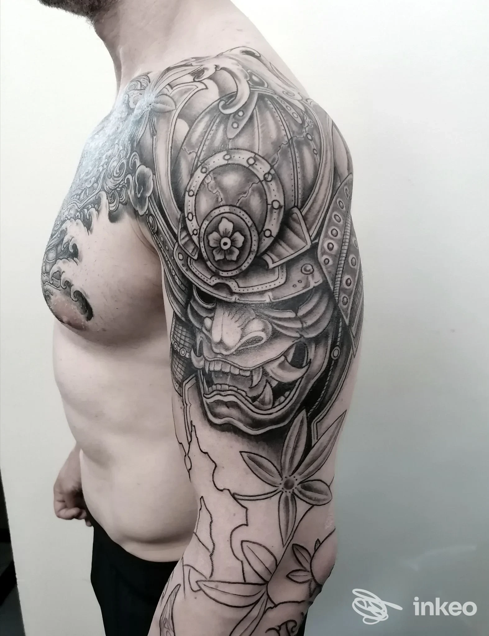 Tattoo a tema meccanico su braccio e spalla, con ingranaggi e piastre dettagliate in bianco e nero; motivi floreali in basso si intrecciano al design principale, su pelle reale.