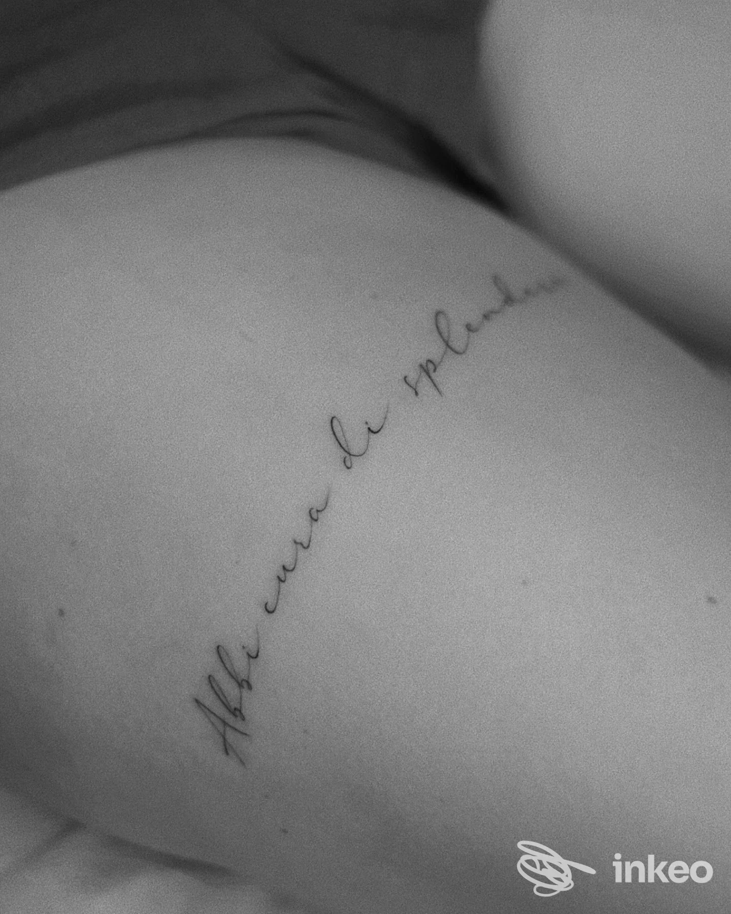 Tattoo in corsivo lungo la spalla di una persona, linea sottile che segue la curvatura della pelle, foto in bianco e nero senza testo o overlay.