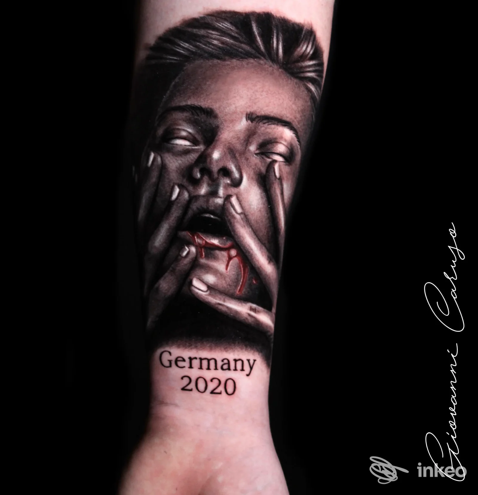 Tattoo realistico su avambraccio raffigurante una faccia con mani al volto e tracce di sangue, con la scritta Germany 2020; realizzato su pelle.