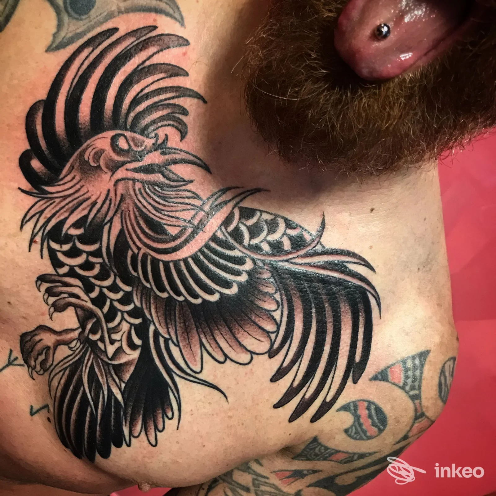 Ampio tatuaggio nero a forma di gallo sul petto con altri tatuaggi sulla spalla; visibile anche una perforazione alla lingua. Miss Machine Tattoo, Montecassiano.