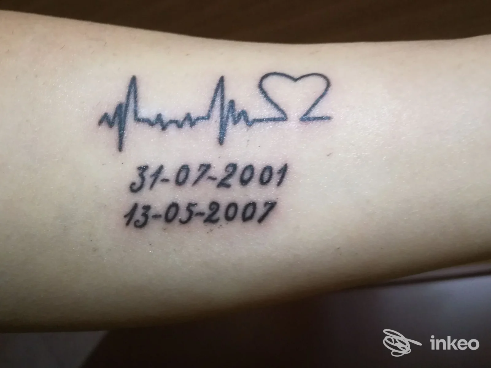 Tattoo sull’avambraccio raffigurante un battito cardiaco seguito da un cuore e due date, senza testo promozionale visibile.