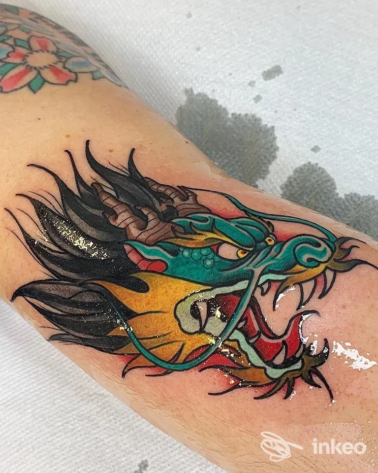 Tatuaggio colorato di un drago su una parte del braccio, linee marcate e riempimenti vividi in tonalità verdi, azzurre e gialle, senza persona visibile.