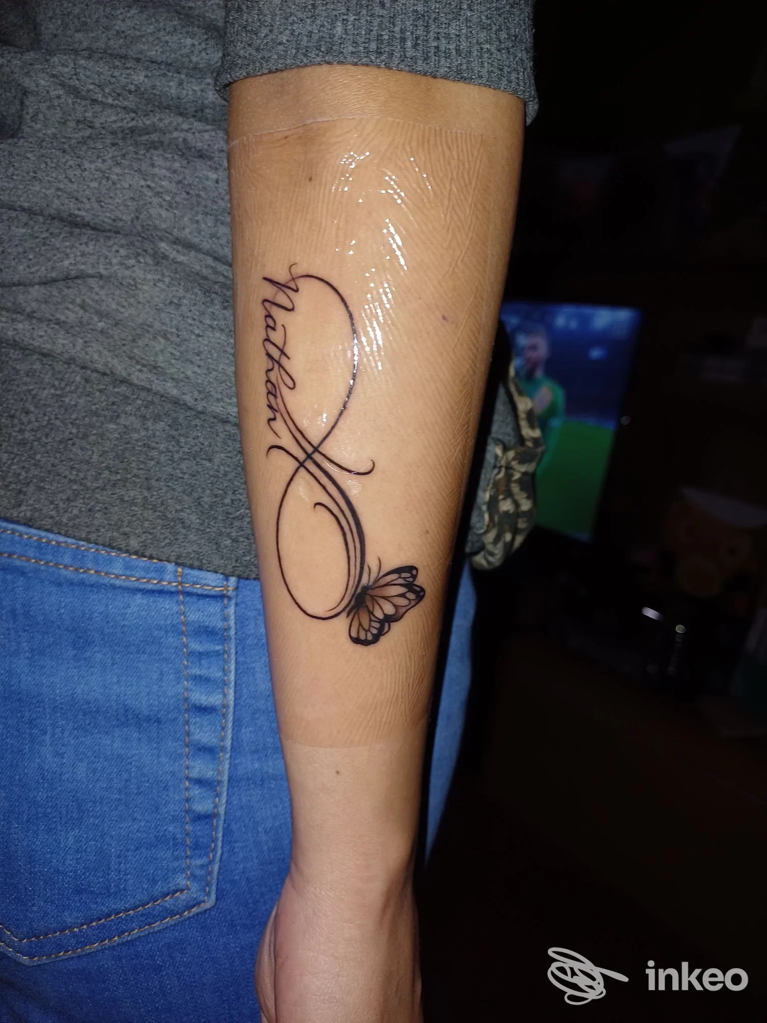 Primo piano di un avambraccio tatuato con scritta calligrafica che sfocia in una piccola farfalla; pelle avvolta in pellicola protettiva — Still Hurts Tattoo Art, Sora.
