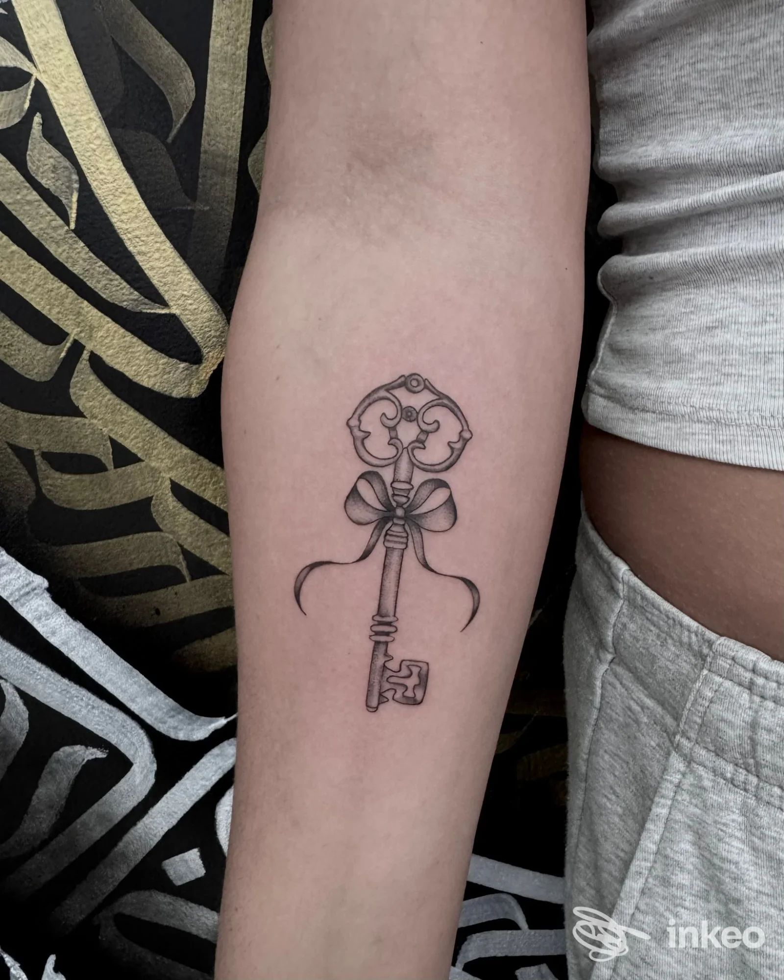 Tatuaggio a inchiostro nero di una chiave decorativa con fiocco sull'avambraccio, realizzato da Black Queen Tattoo a Oderzo.