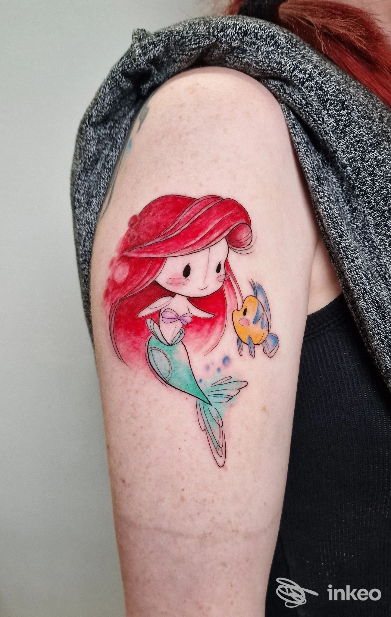 Tatuaggio colorato sul braccio superiore con una sirenetta in stile cartoon e un piccolo pesce, linee sottili e colori vividi. Ludwig Tattoo, Bologna.
