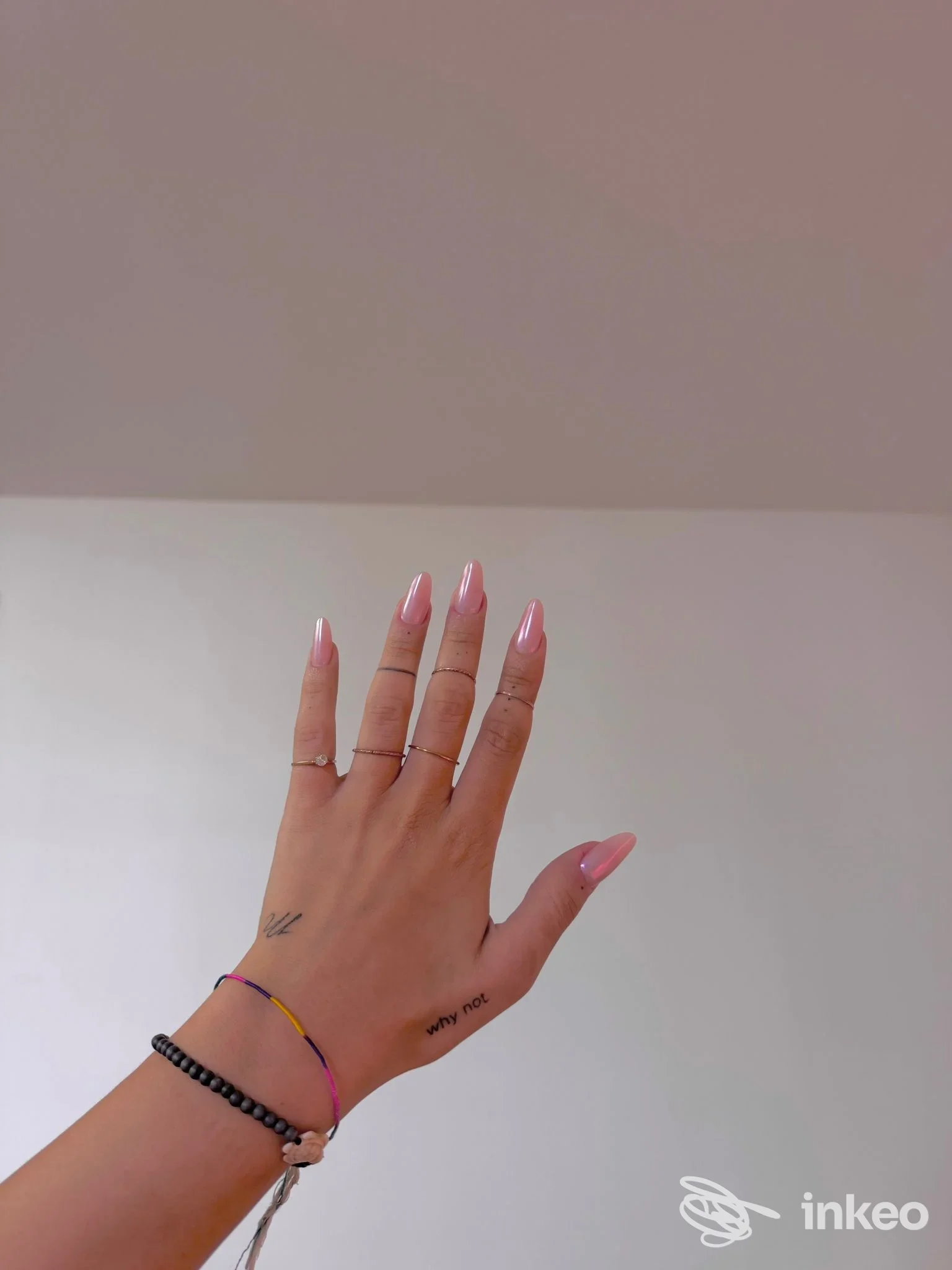 Mano con piccoli tatuaggi su polso e lato della mano e smalto rosa su sfondo neutro. Perla Tattoo Studio, Bologna.