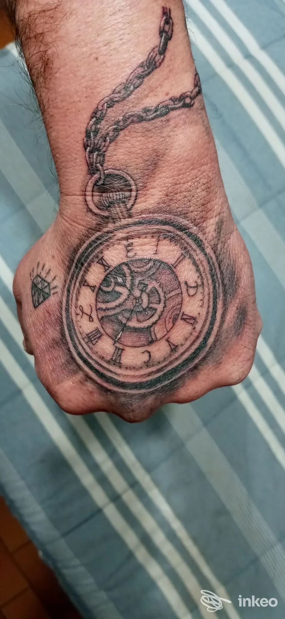 Tatuaggio di orologio da tasca con catena sul dorso della mano, piccolo simbolo a diamante. Black Wave Studio, Pietrasanta.