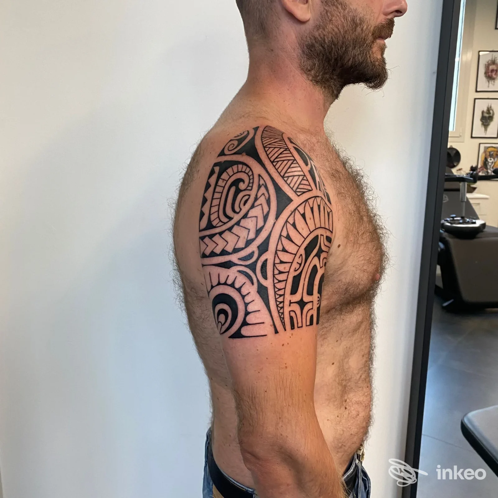 Braccio di uomo con tatuaggio geometrico nero dall’omero alla spalla, viso parzialmente visibile, in studio di tatuaggi.