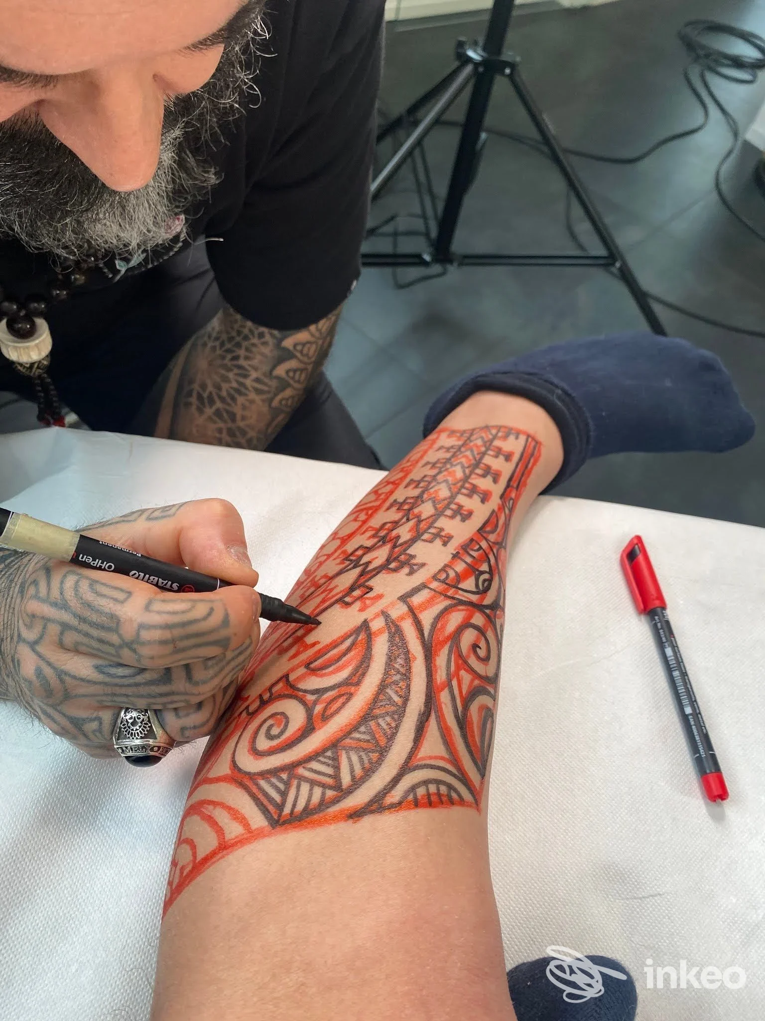 Un tatuatore lavora sull'avambraccio di un cliente, disegnando linee rosse geometriche su pelle nuda, con strumenti di disegno visibili sul tavolo. Nessun volto visibile.