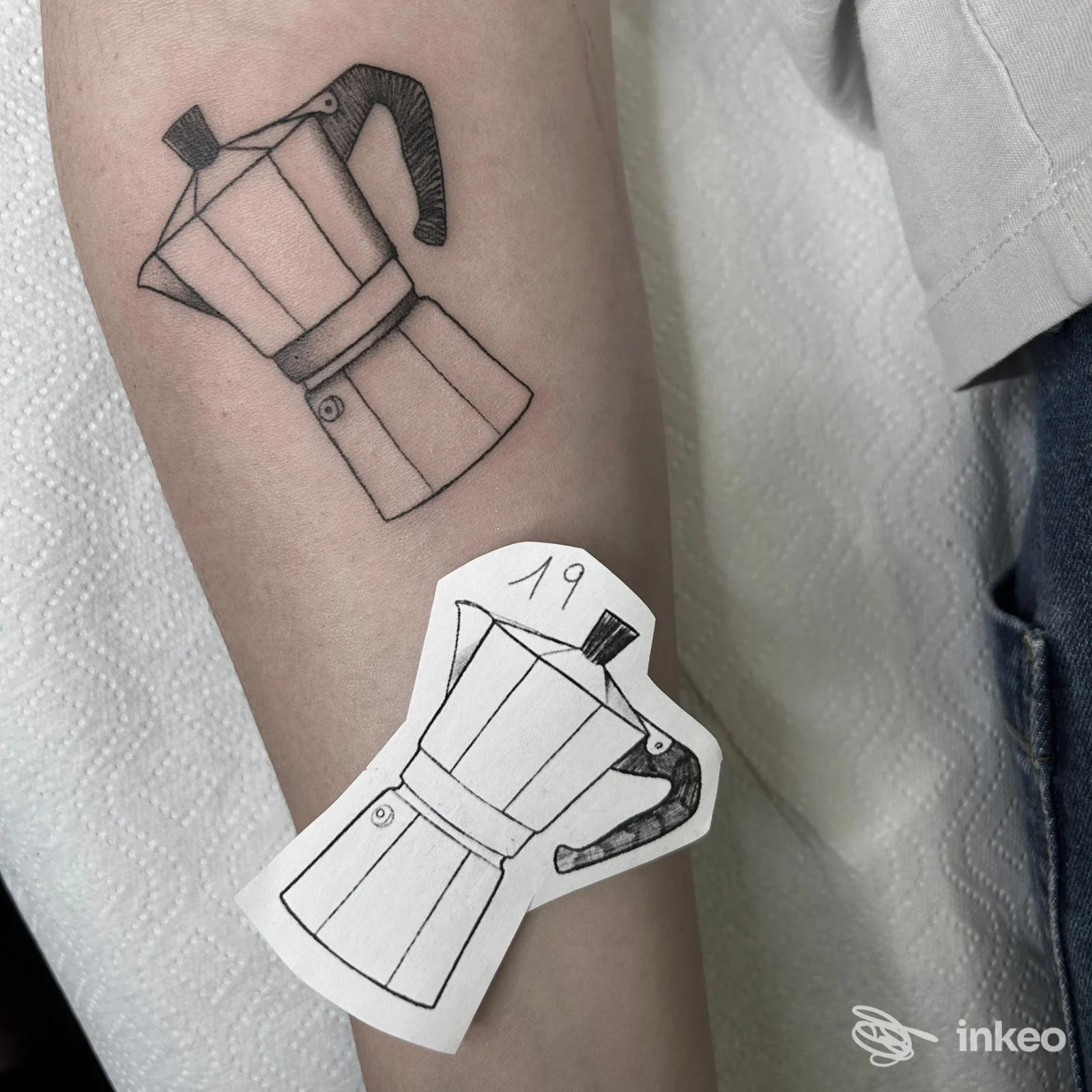 Tatuaggio di una caffettiera moka in linework sull'avambraccio, con il bozzetto su carta accanto. Studio Matita Tattoo, Bergamo.