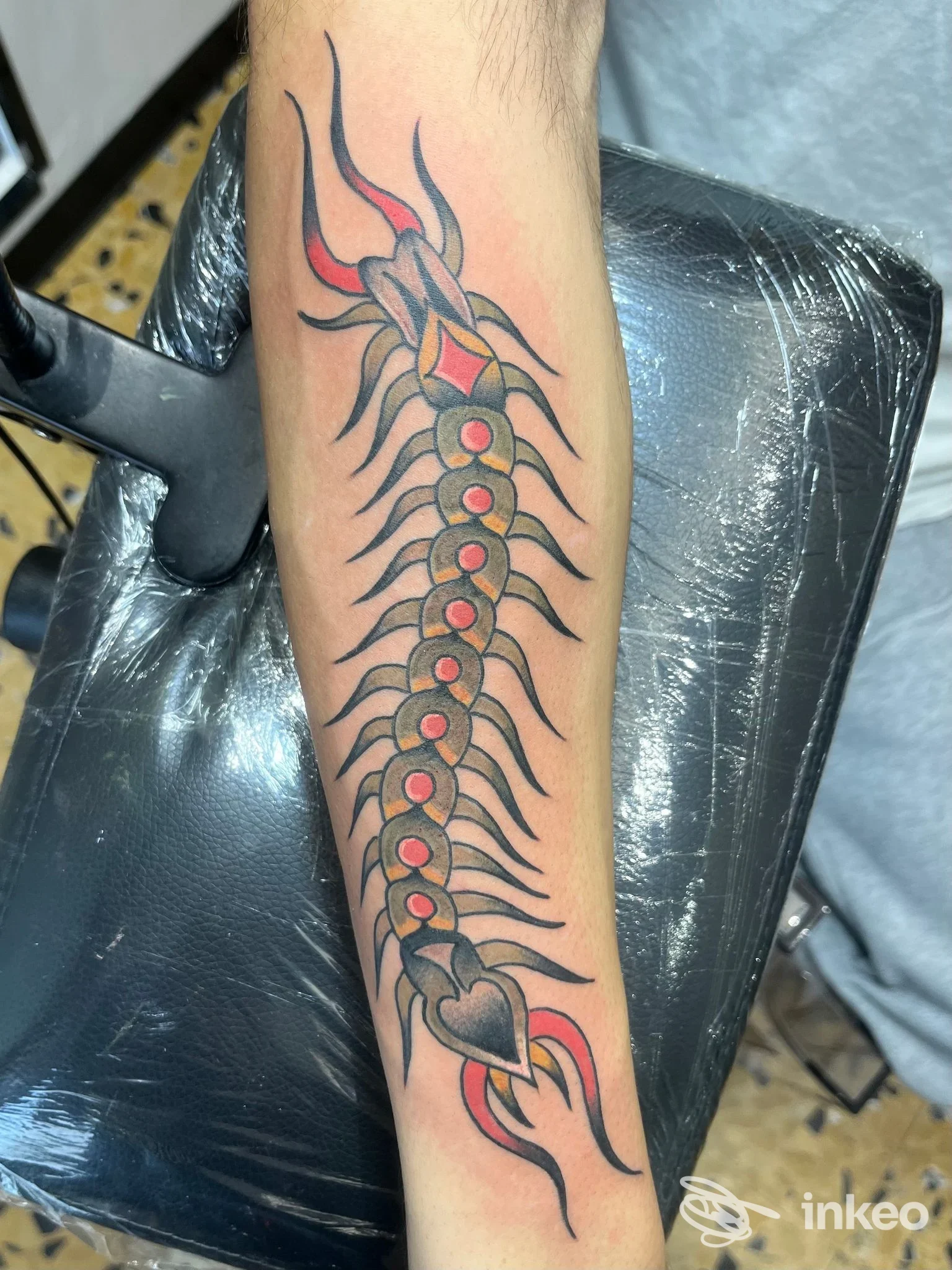 Tatuaggio lungo il braccio che simula una colonna vertebrale con costole stilizzate e un cuore in basso, con dettagli rossi e linee nere, realizzato direttamente sulla pelle.